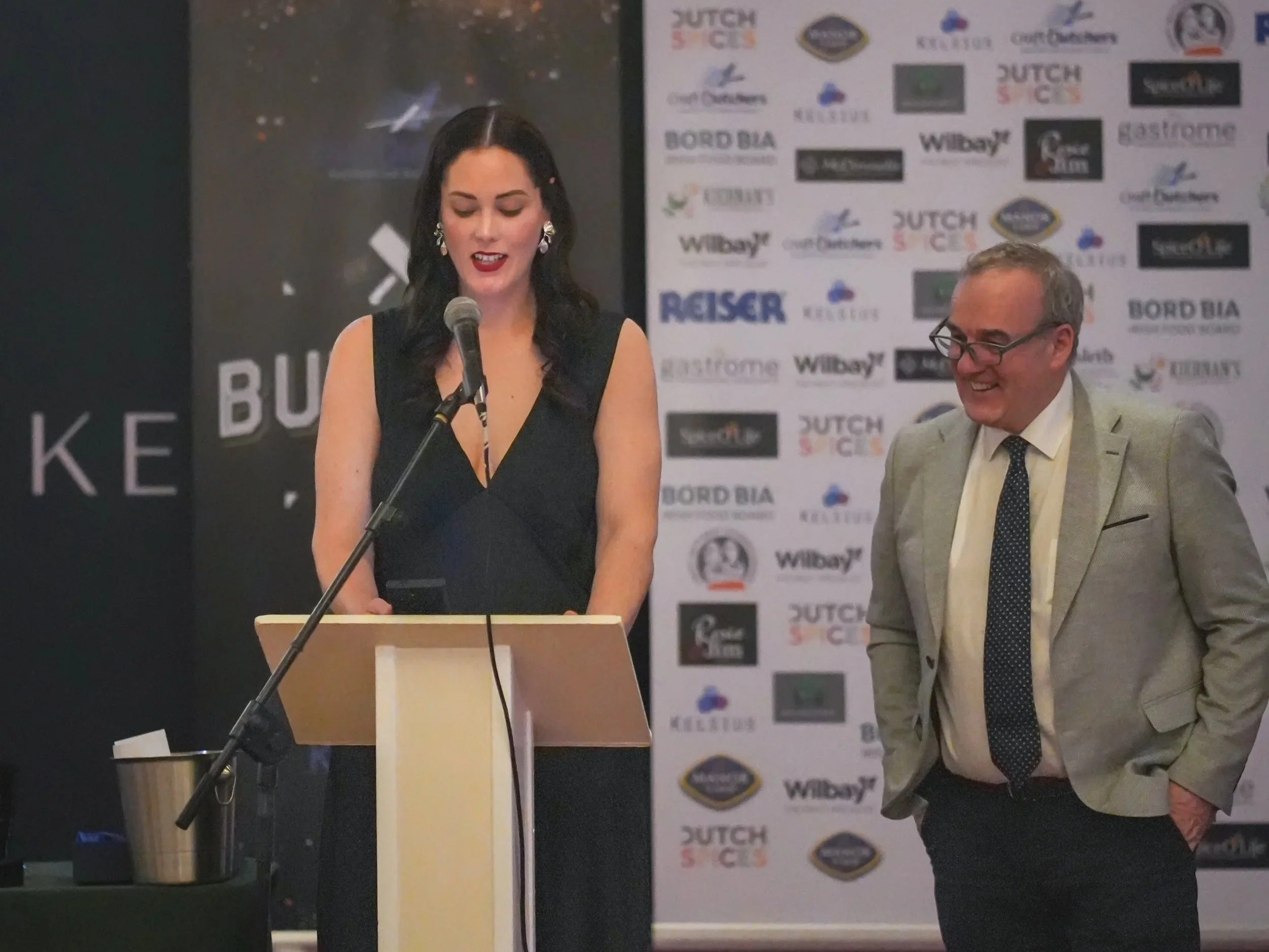 Alayna the Kiwi Butcher is speaking at a podium with a microphone, wearing a black dress and earrings, with a background that has various sponsor logos and a man sitting beside her, smiling, dressed in a beige suit and dark tie.