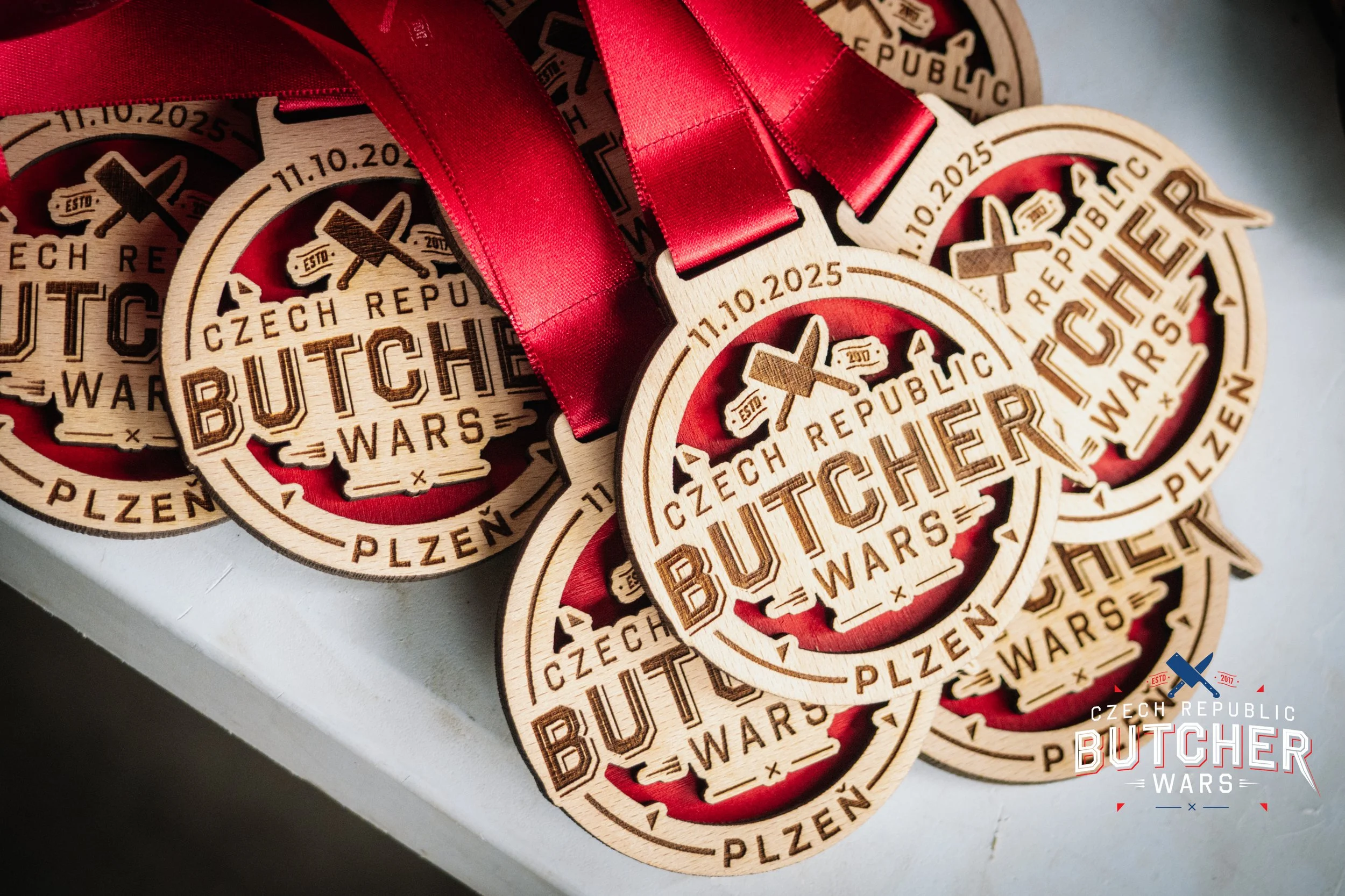 Multiple wooden medals with red ribbons, engraved with text 'Czech Republic Butcher Wars Plzeň 11.10.2025' and a graphic of a cleaver and fork.