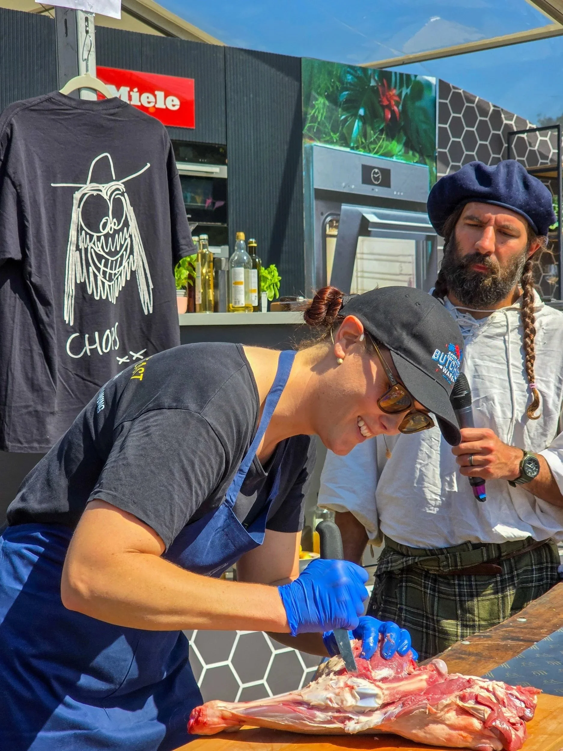 A woman wearing a black cap, glasses, and blue gloves is cutting raw meat on a wooden surface at an outdoor food event. A man with a beard, braids, and a blue beret, holding a microphone, observes. Behind them are hanging shirts, bottles, and a grill in the background.