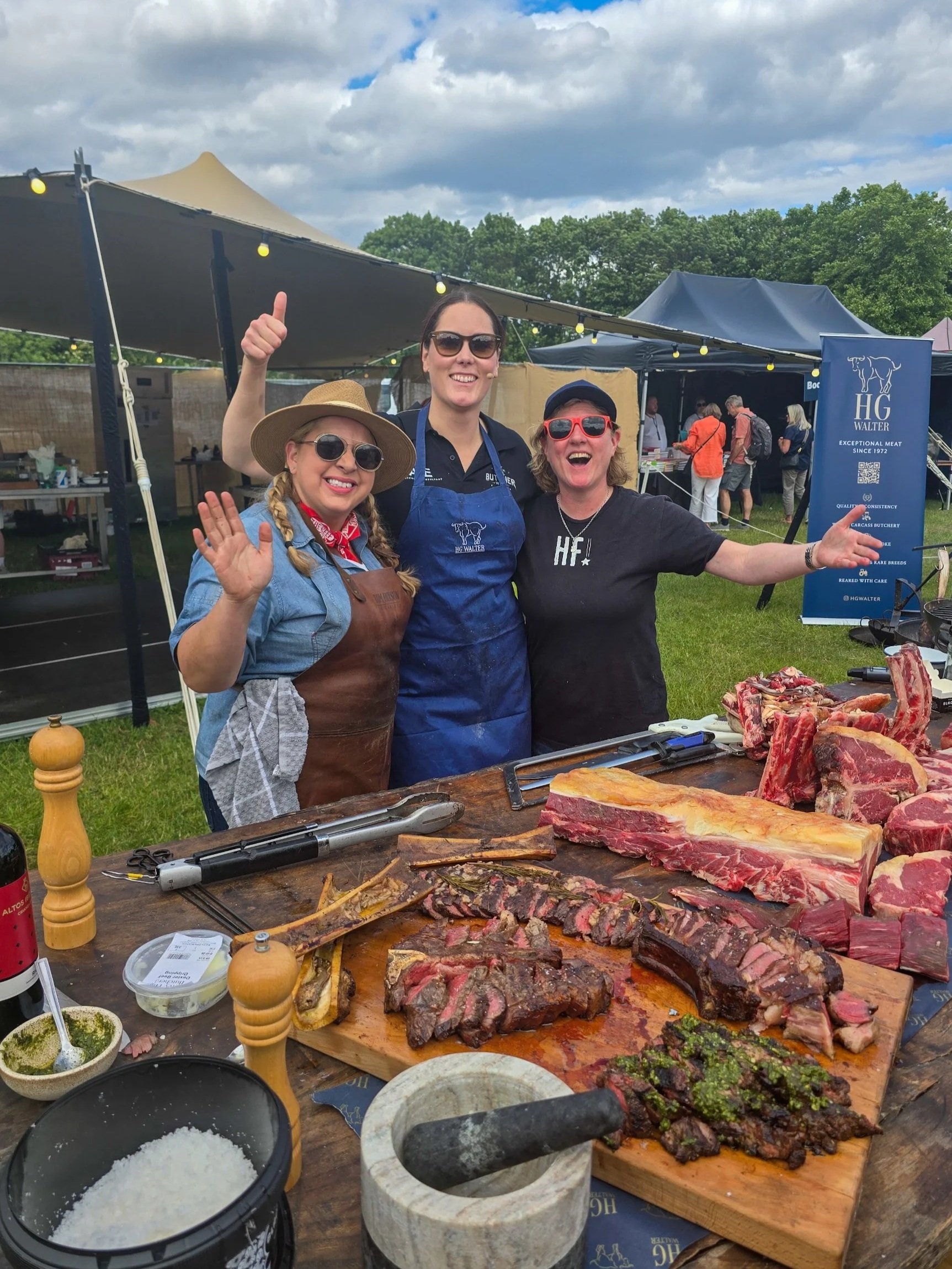 Sam Evans, Alayna Empson and Shauna Guinn waving at a barbecue event, standing behind a table with various cuts of expertly cut dry aged beef, salt, pepper, and condiments, outdoors under cloudy skies at Pub in the Park London in 2025.