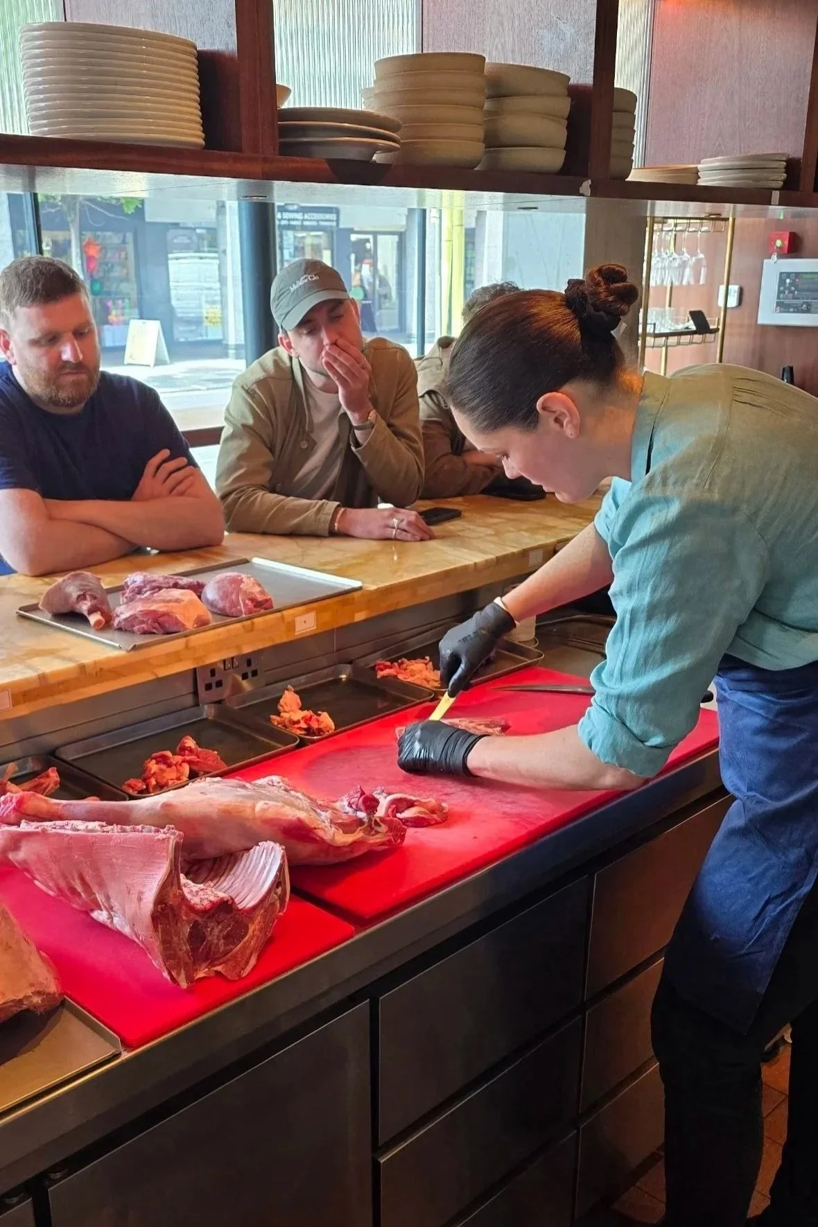 A butcher wearing black gloves slicing lamb on a red cutting board, with chefs watching in a restaurant or butcher shop. There are plates on shelves above, and a large window in the background.