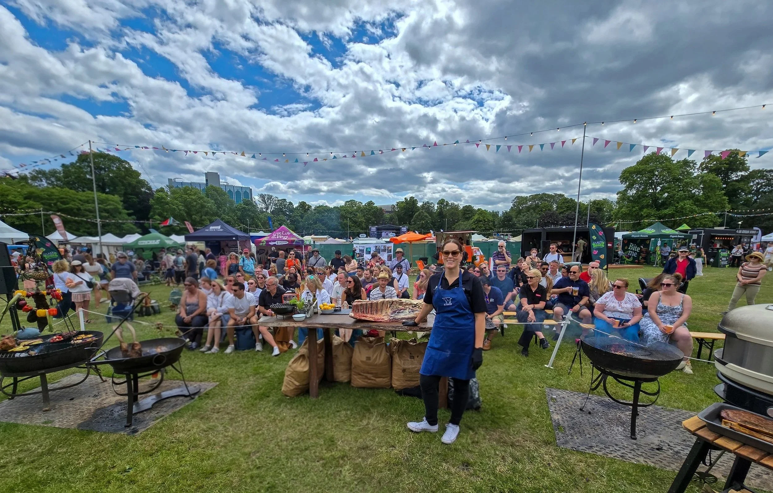 A large outdoor festival with a crowd of people seated on benches on grassy field, several food trucks and tents in the background, a woman smiling in the foreground with a blue apron in front of cooking grills, under a partly cloudy sky with colorful bunting overhead.