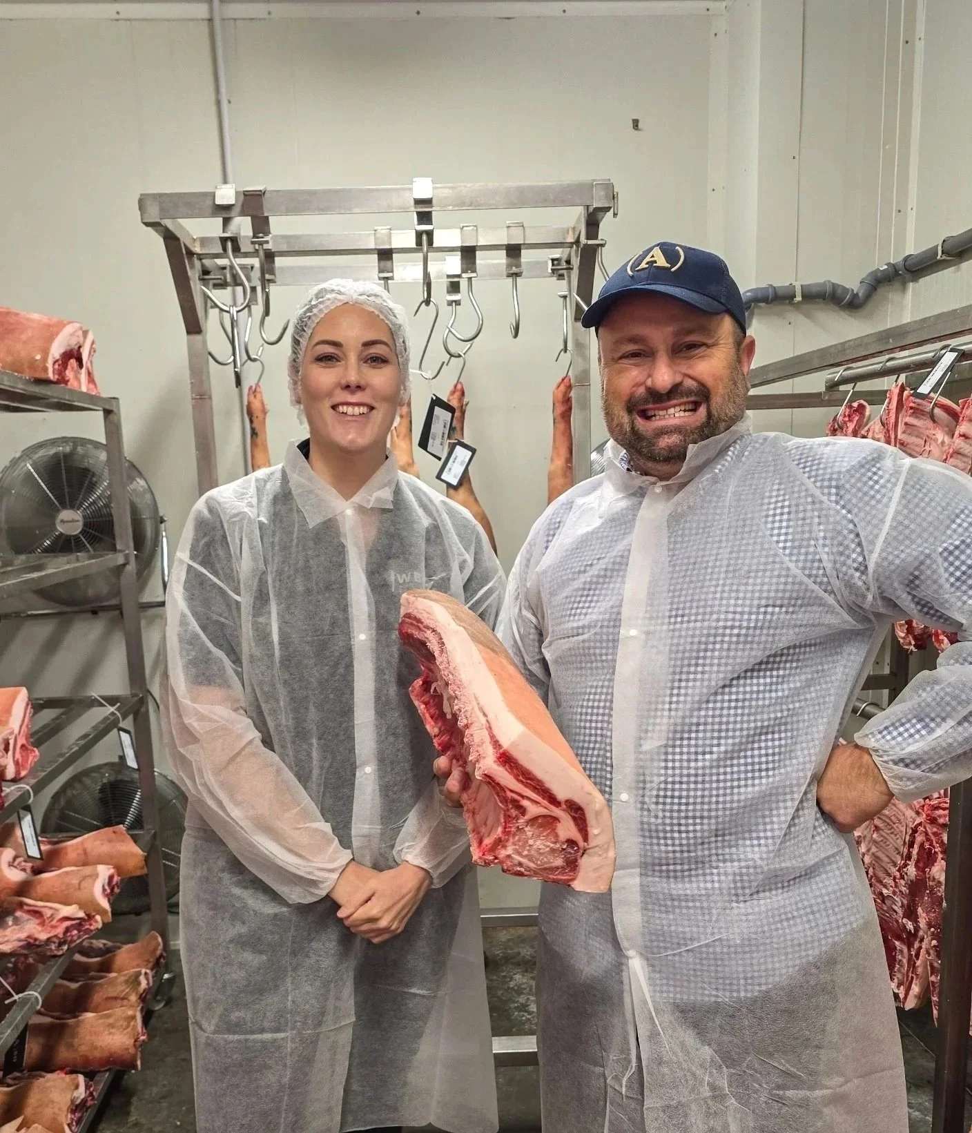 Two people in a meat processing facility holding a large cut of meat, with shelves of meat in the background and hooks hanging from a metal rack.