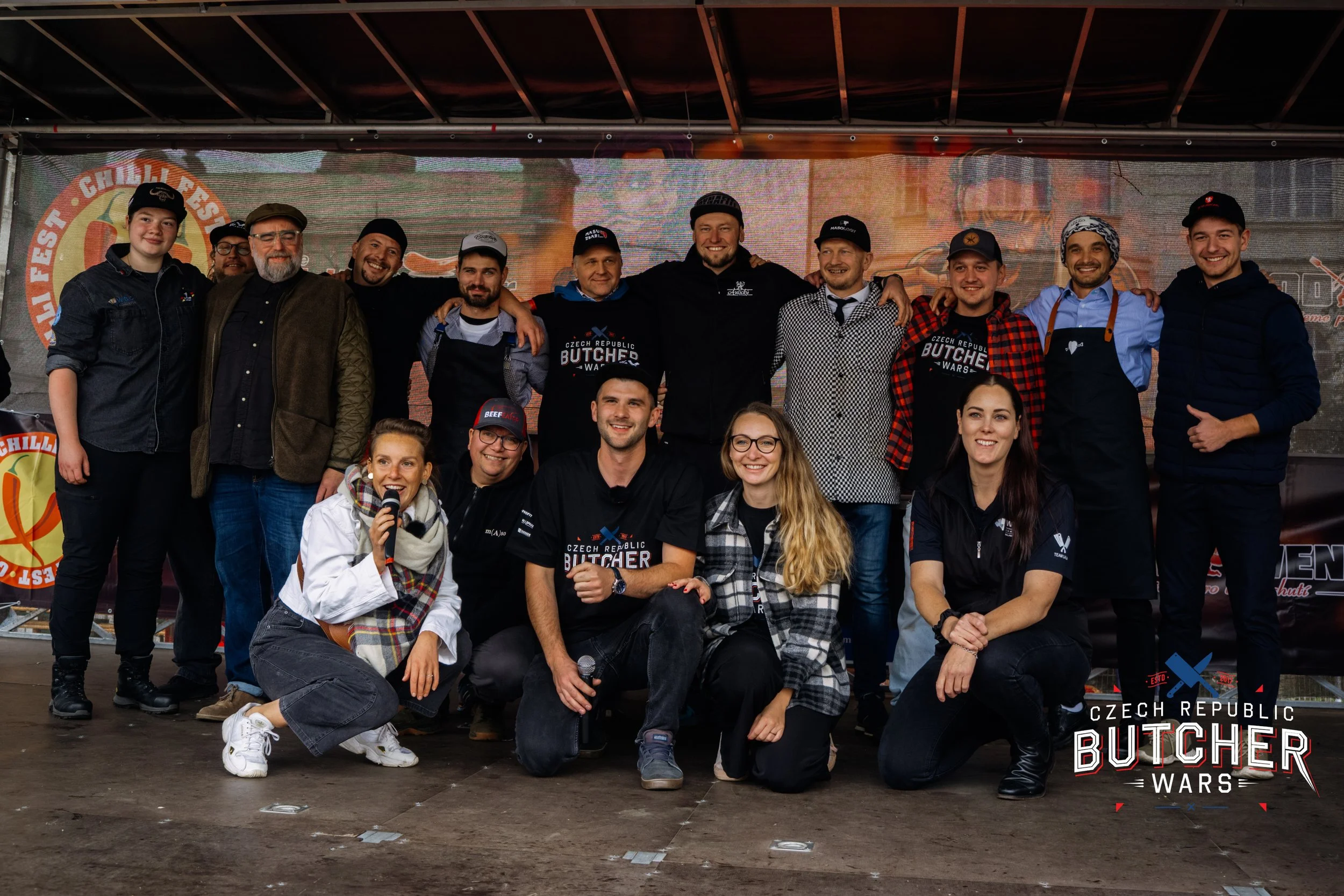 A group of fifteen people posing together on a stage at a Czech Republic butchery festival, with some holding a microphone and others smiling, wearing casual and chef attire, with a banner in the background displaying the event's logo.