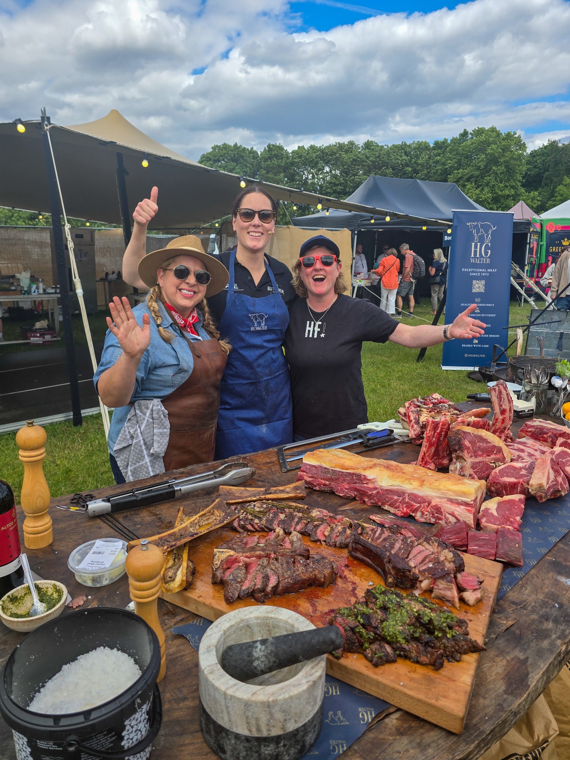 Sam Evans, Alayna Empson and Shauna Guinn waving at a barbecue event, standing behind a table with various cuts of expertly cut dry aged beef, salt, pepper, and condiments, outdoors under cloudy skies at Pub in the Park London in 2025.