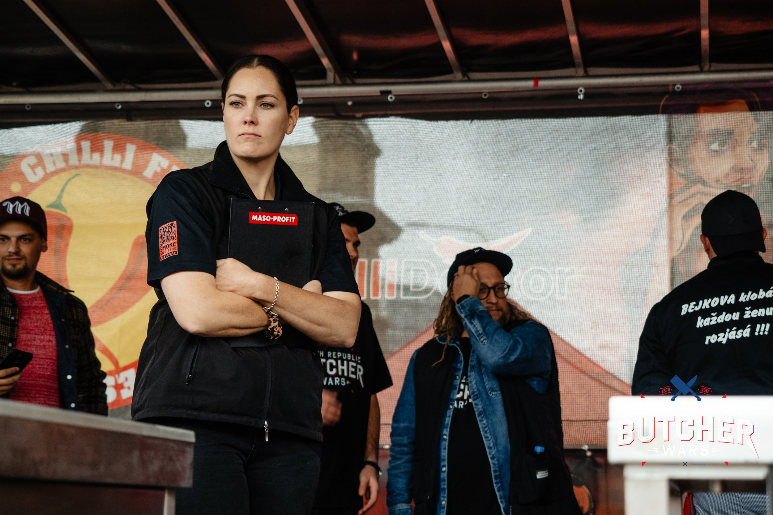 Butcher & Judge Alana Empson dark hair wearing a black jacket crossing her arms on a stage, surrounded by several other people, some wearing black and denim jackets, with a large illuminated sign that reads 'BUTCHER WARS' in the background.