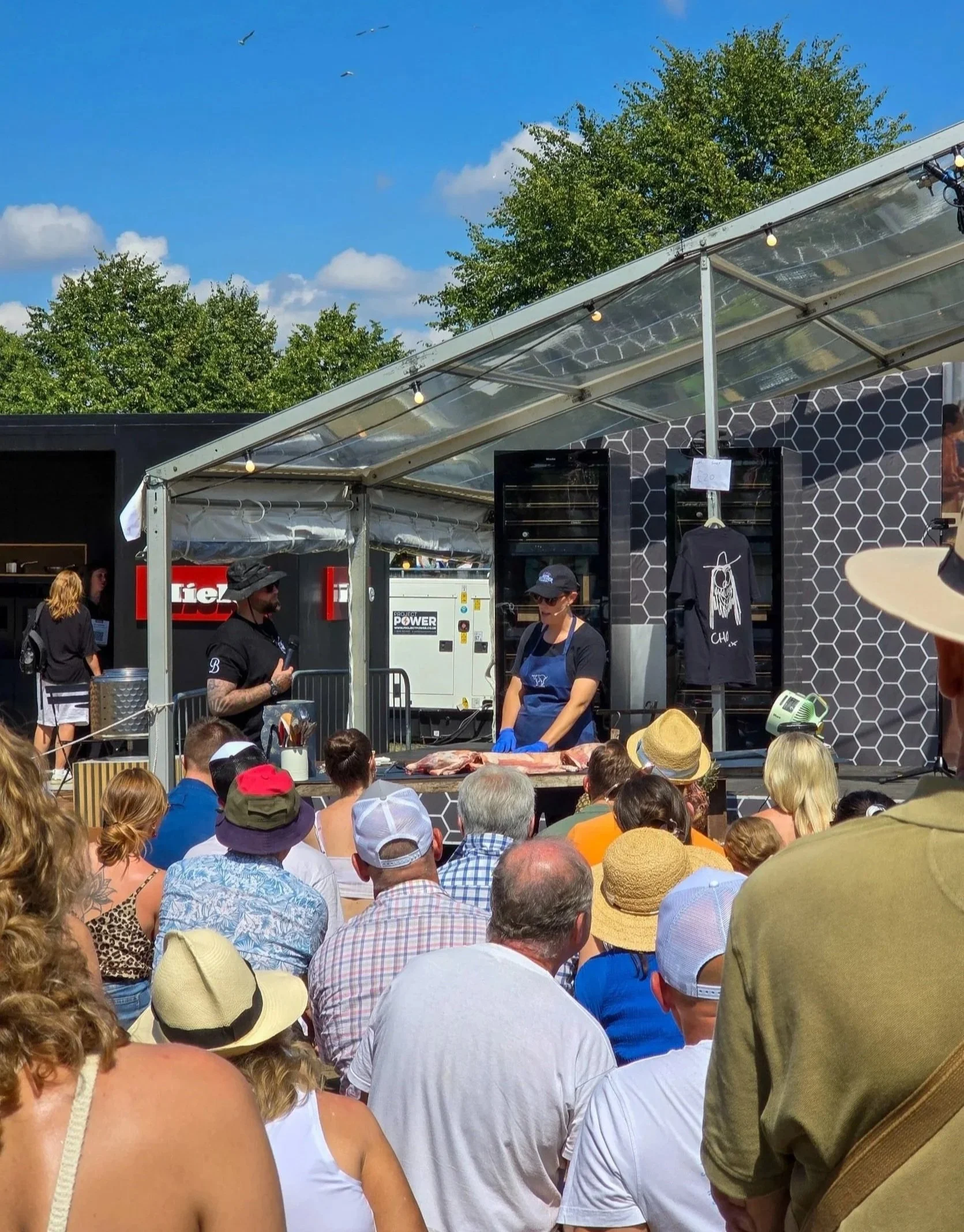 Crowd attending a cooking demonstration outdoors with a chef preparing meat, under a partly cloudy sky and surrounded by green trees.