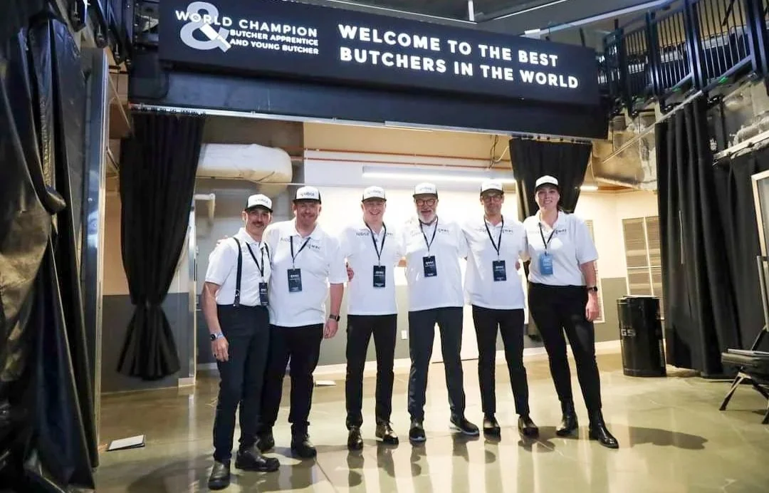Group of six expert butchery judges wearing white shirts, black pants, and white caps, standing under a sign that reads 'Welcome to the best Butchers in the World' at the Golden One Centre in Sacramento California ahead of the WBC in 2022.