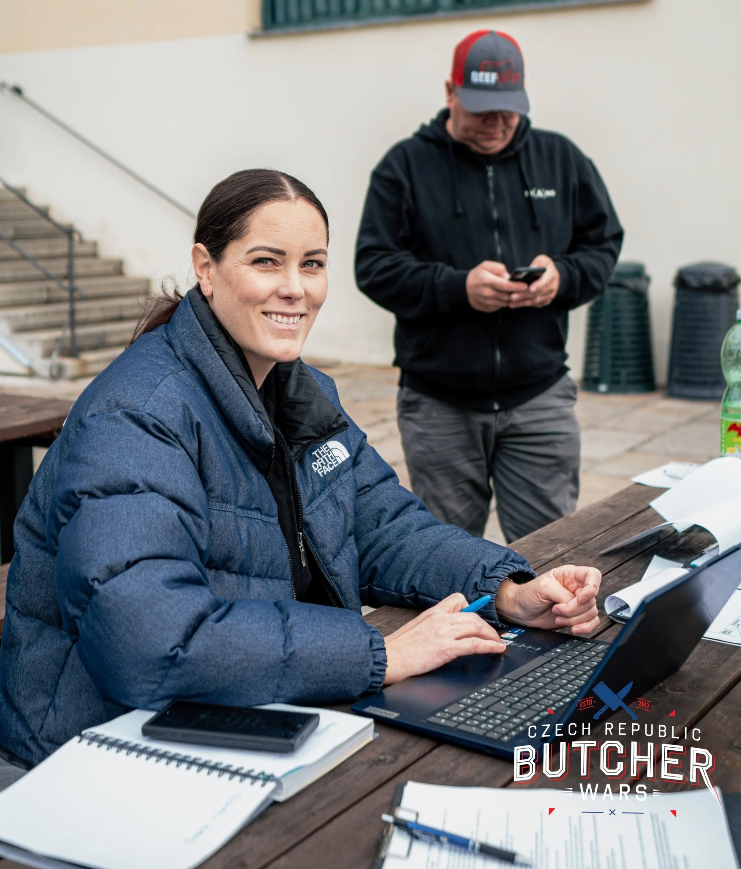 A woman in a blue jacket sitting at a wooden table with a laptop, notebook, and smartphone, smiling at the camera. A man in a black hoodie and cap stands behind her, looking at his phone. The table has papers and a bottle of soda. The bottom right corner has a logo that reads 'Czech Republic Butcher Wars.'