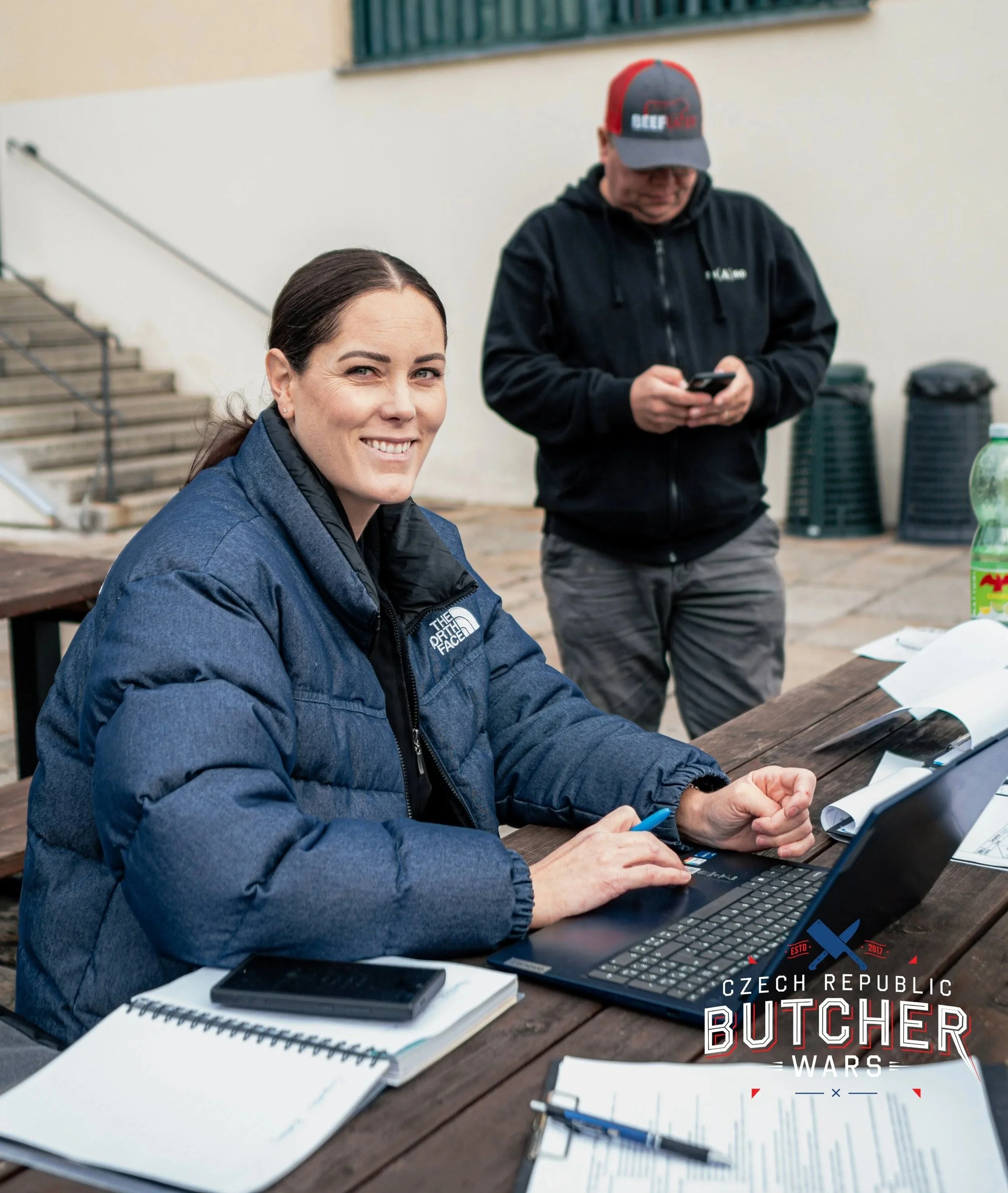 A woman sitting at a wooden table with a laptop is smiling at the camera. She is wearing a blue North Face jacket. There are notebooks, papers, and a smartphone on the table. In the background, a man is standing and looking at his phone. The scene takes place outdoors with steps and a building in the background. There is a logo at the bottom right that reads 'Czech Republic Butcher Wars'.