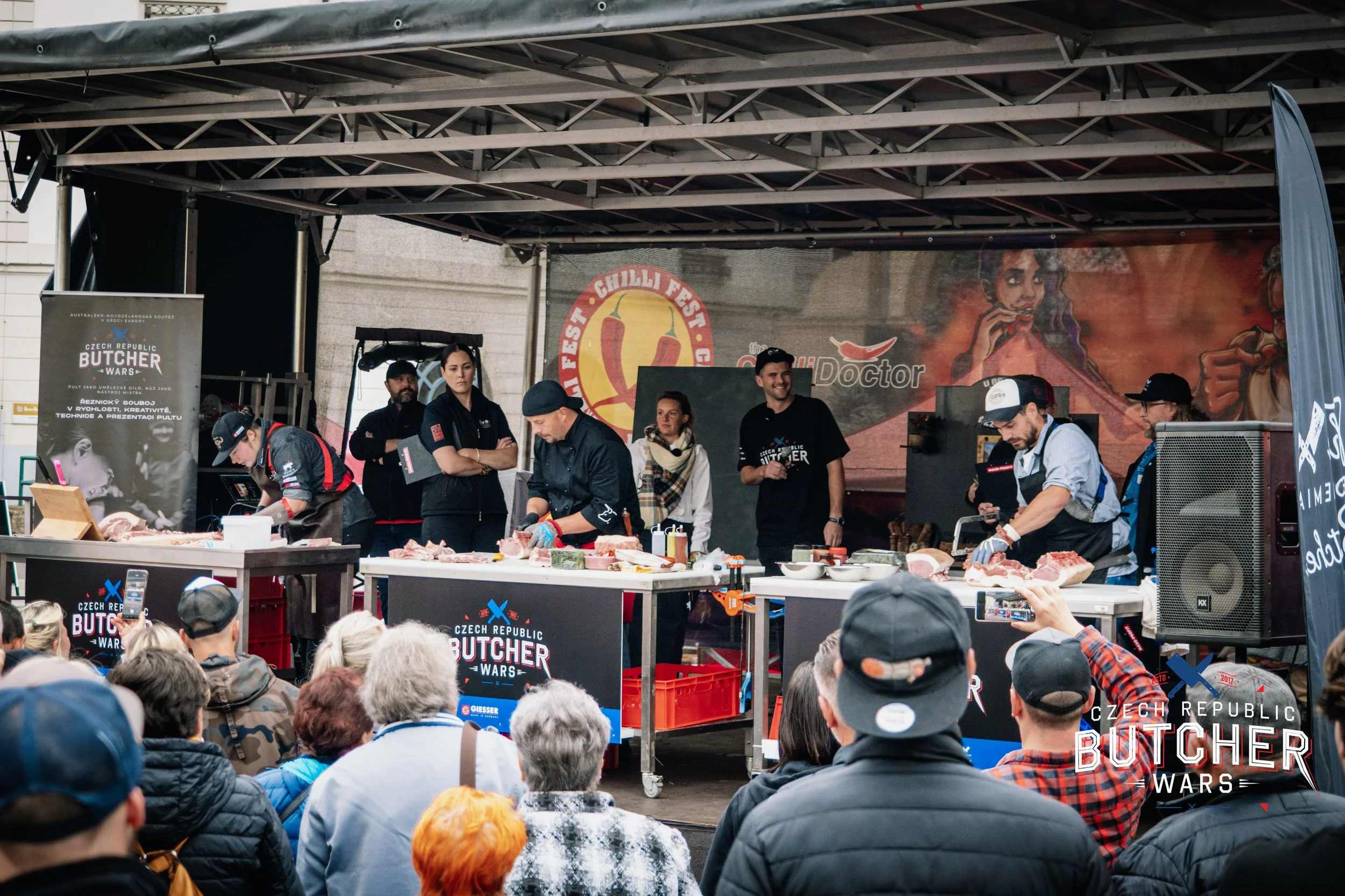 A cooking competition stage with buchers preparing meat in front of a crowd at the Czech Republic Butcher Wars event, with several judges observing and a large banner in the background.
