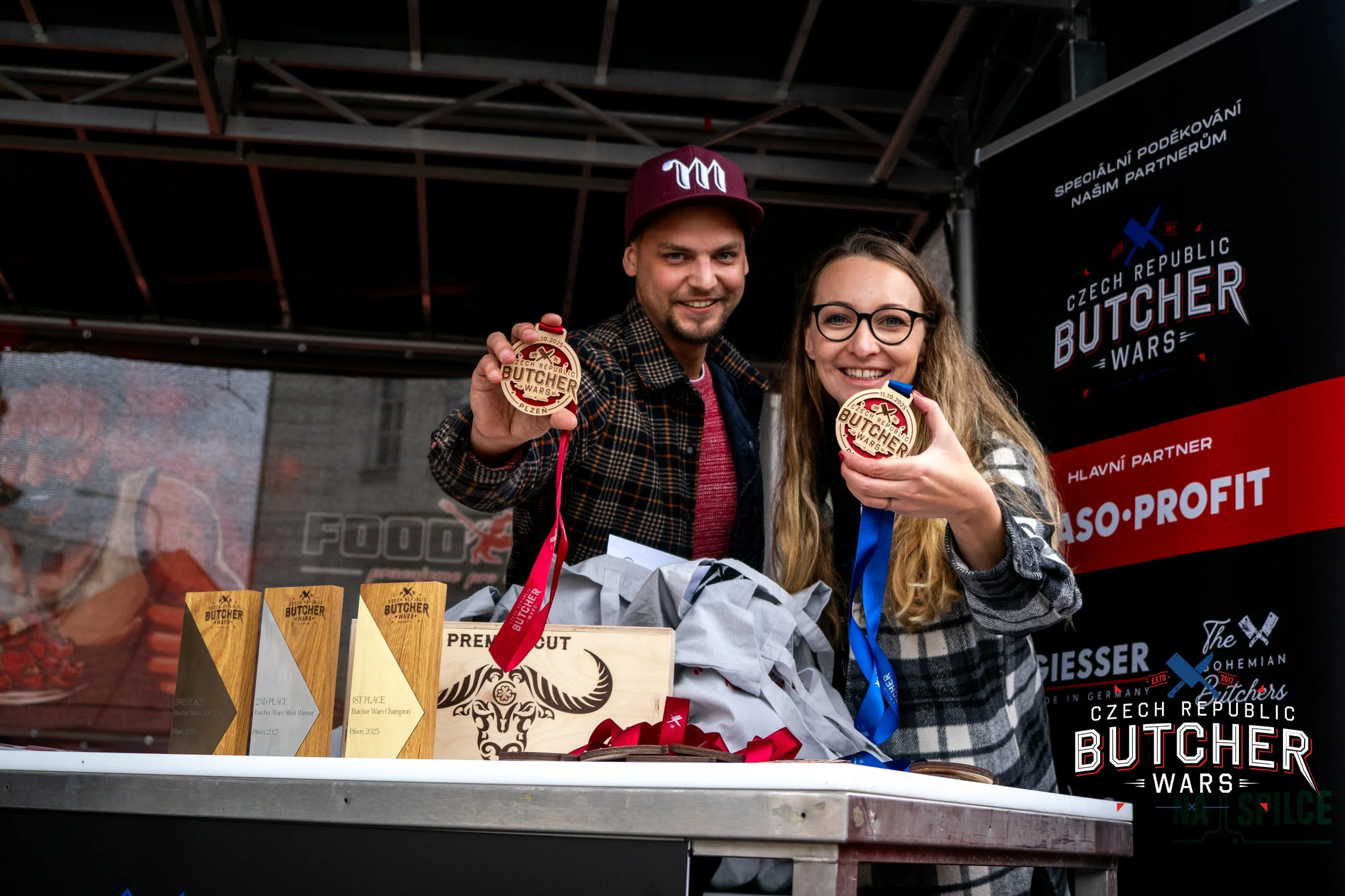 Two people, a man and a woman, smiling and holding medals at the Czech Republic Butcher Wars event, with trophies and event materials on the table in front of them.