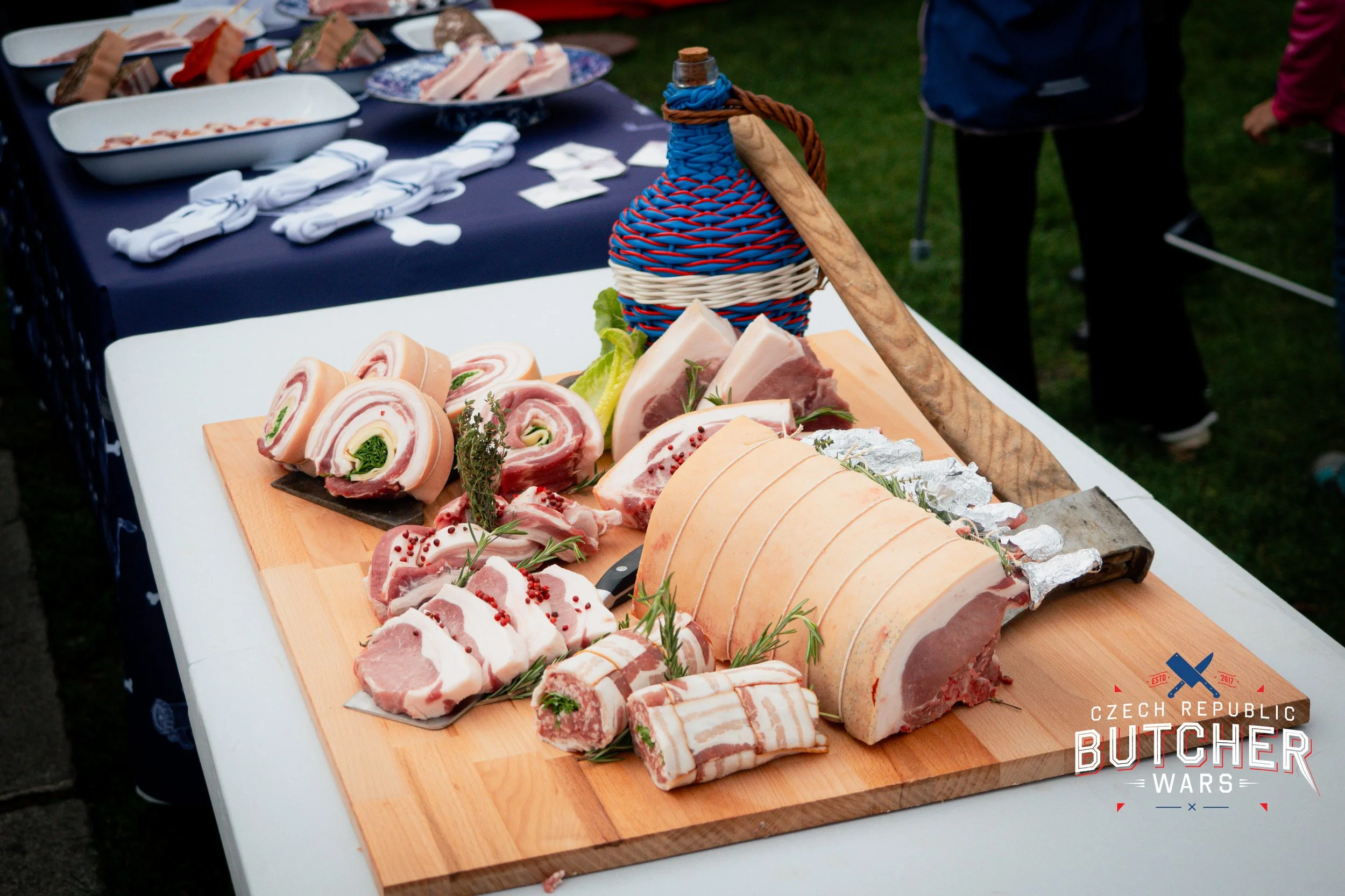 Various cuts of raw pork and meat products on a wooden cutting board at a butcher event, with a bottle wrapped in colorful woven covering and a large knife, at an outdoor setting.