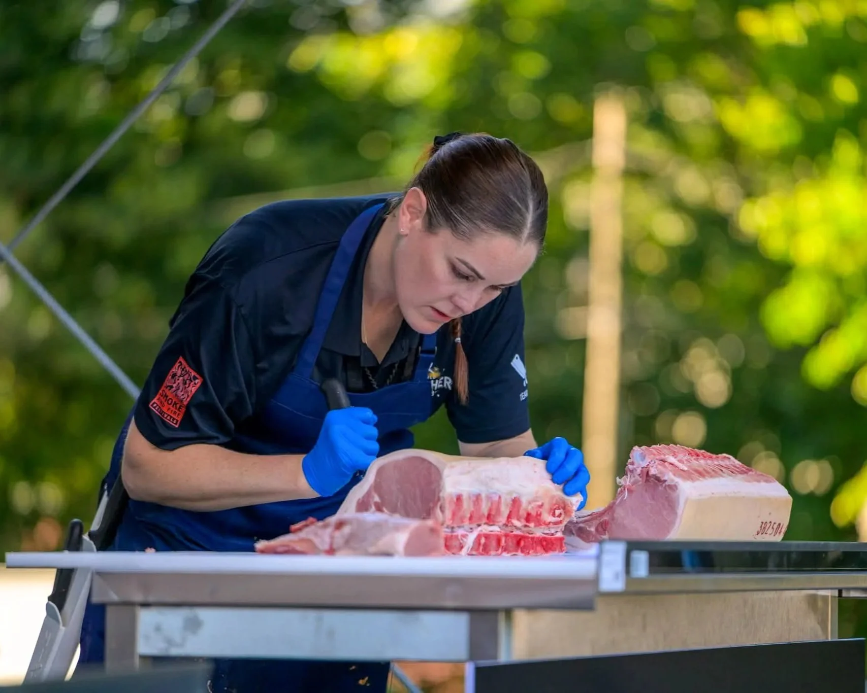 A woman in a black shirt and blue apron expertly cuts a large piece of pork loin on a table outdoors at Transylvania Butcher Wars in 2025.