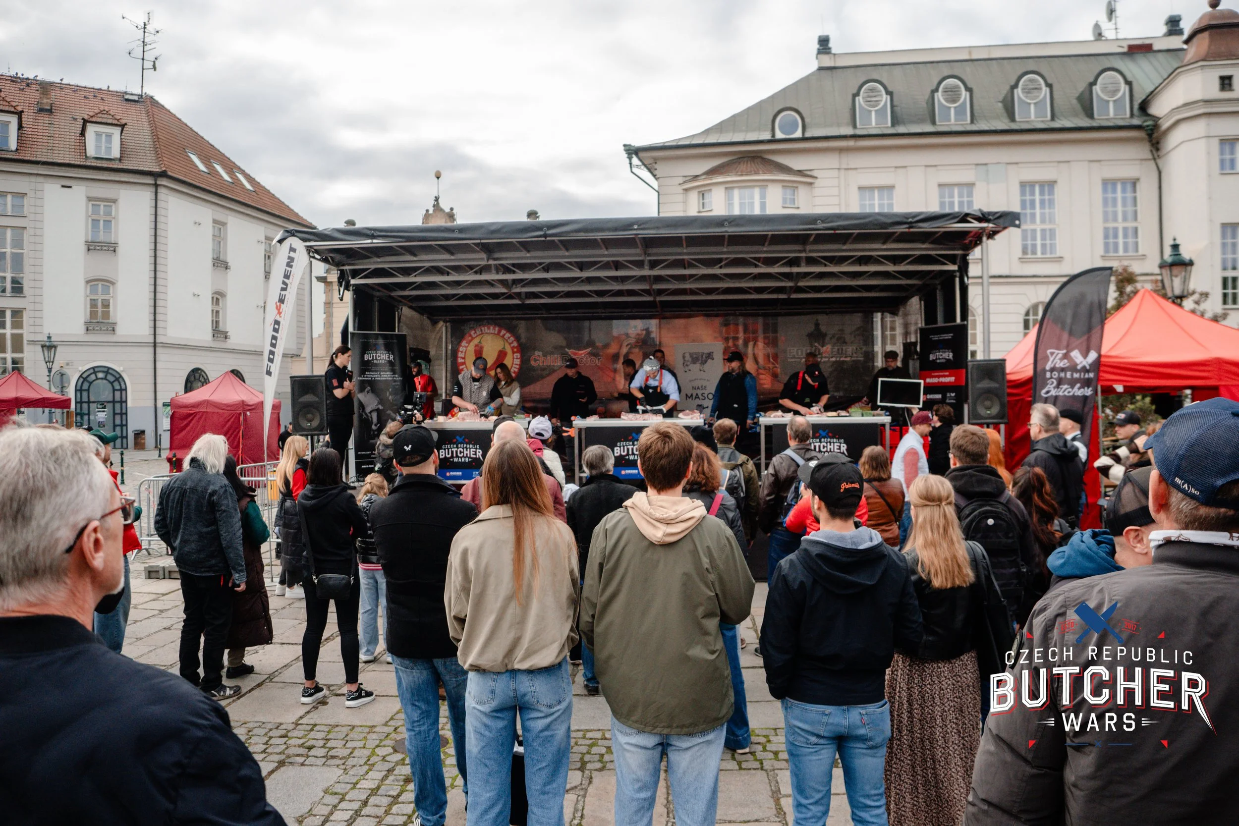 People gathered at an outdoor event watching a stage where multiple chefs are preparing food during the Czech Republic Butcher Wars, with event banners and tents in the background.