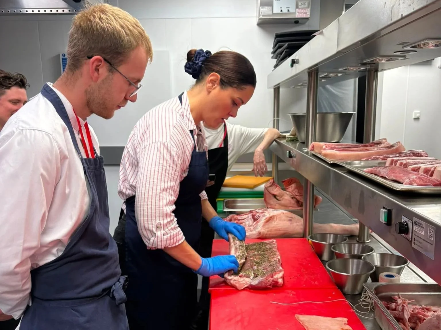 People in a commercial kitchen butchering and preparing meat, with several trays of meat and kitchen tools.