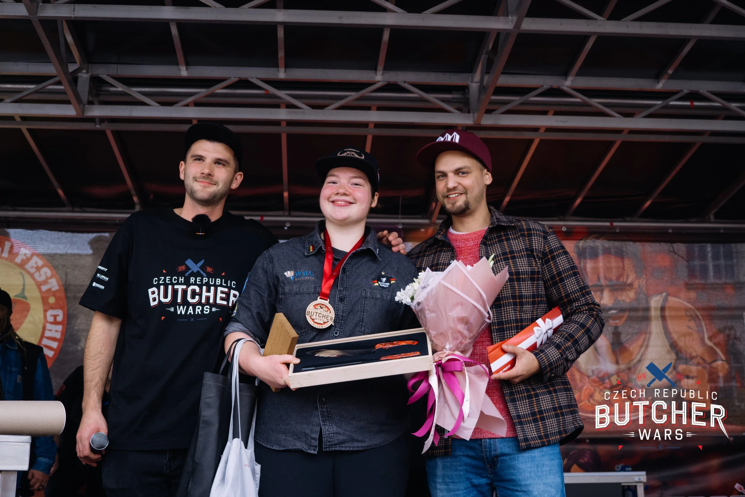 Three young people on stage at a competition event, one holds a gift box, another has a medal, and the third is holding a bouquet and a rolled certificate, with a banner that reads "Czech Republic Butcher Wars" in the background.