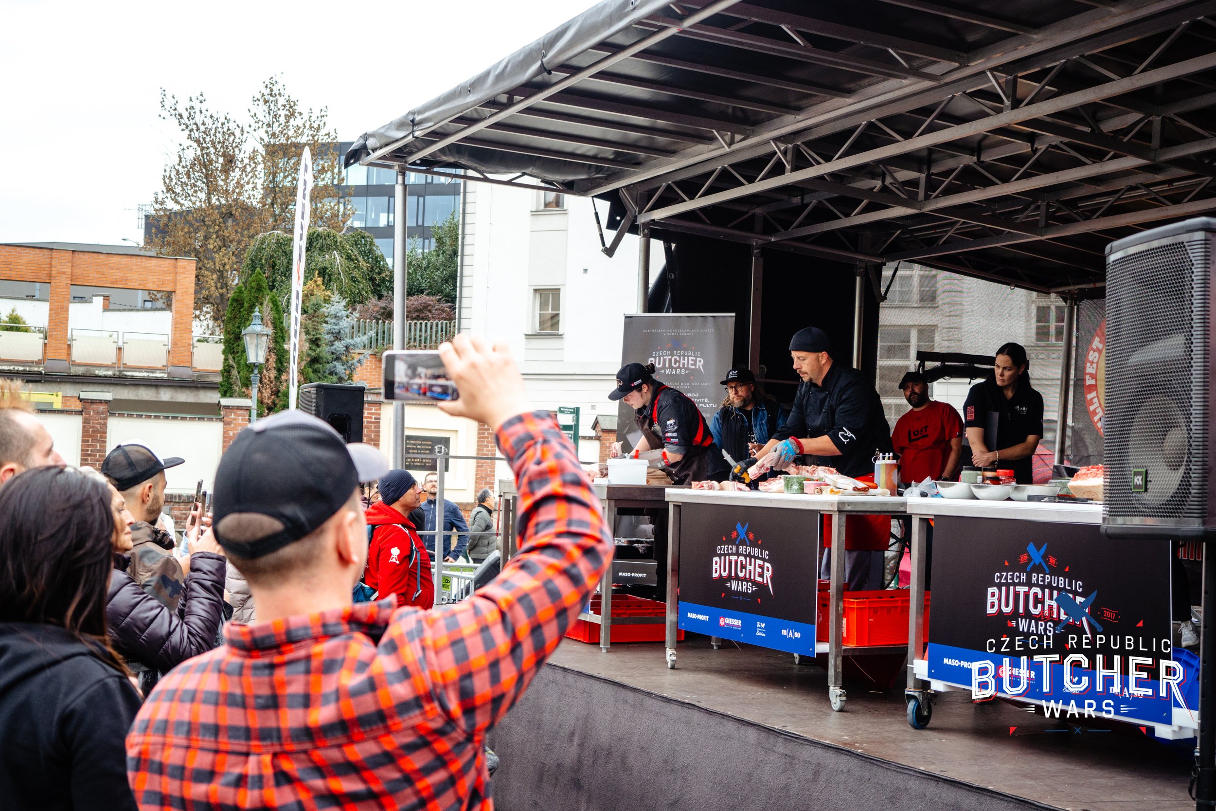 Butchers preparing meat at a culinary event on an outdoor stage with a crowd watching, some taking photos, in front of banners that say 'Czech Republic Butcher Wars'.