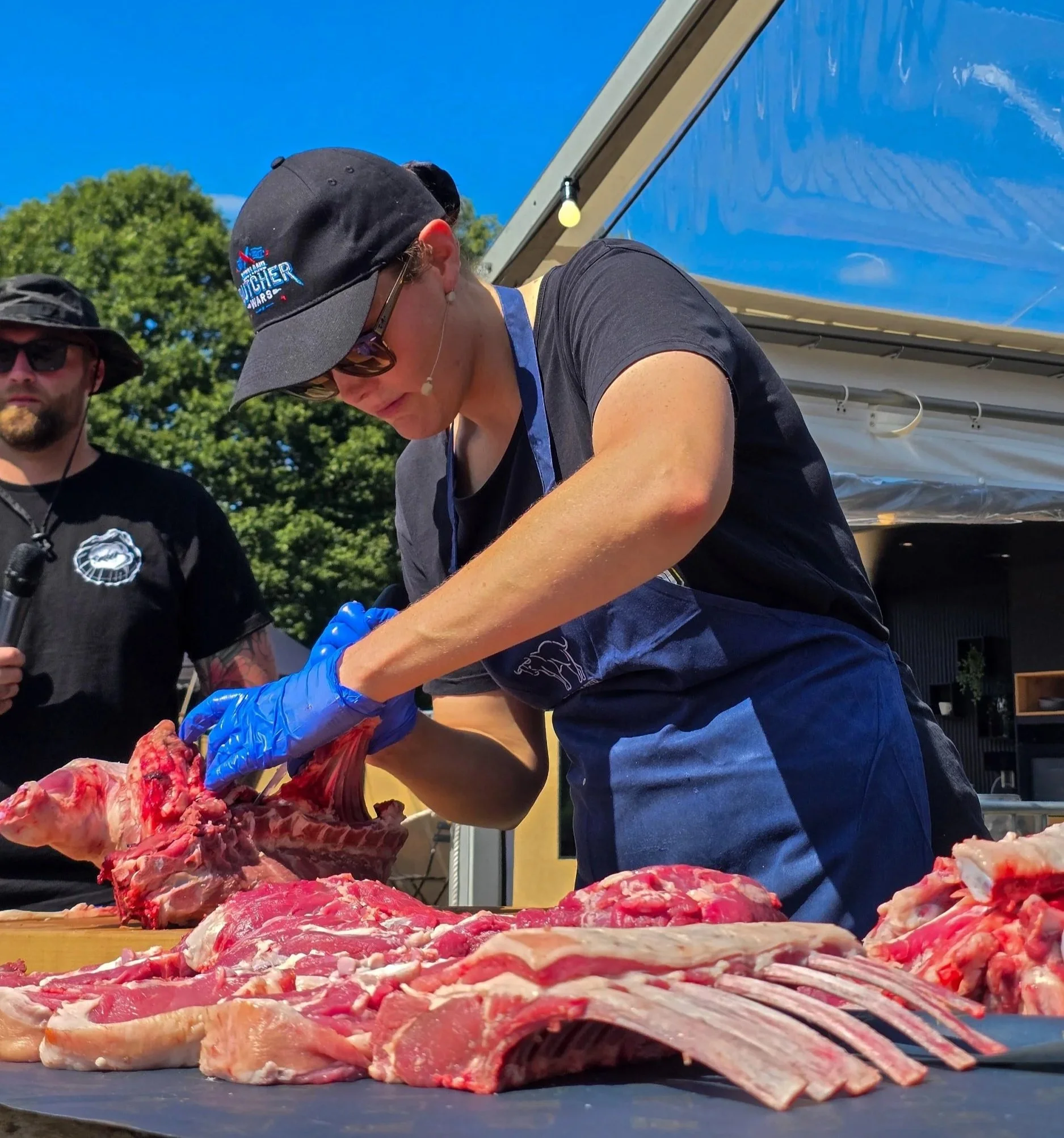 Butcher Alana Empson wearing sunglasses, a black hat, and a black t-shirt while she butchers a side of lamb, outside at Pub in the Park Reigate, with chef James Peck in the background.