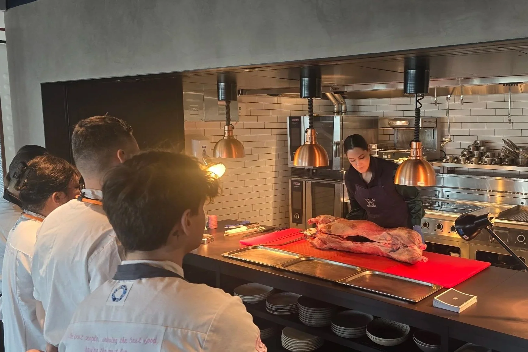 A group of people watching a chef demonstrate butchering a large piece of meat in a modern kitchen setting. The chef is leaning over the meat on a red cutting board, with several pans and kitchen appliances in the background.