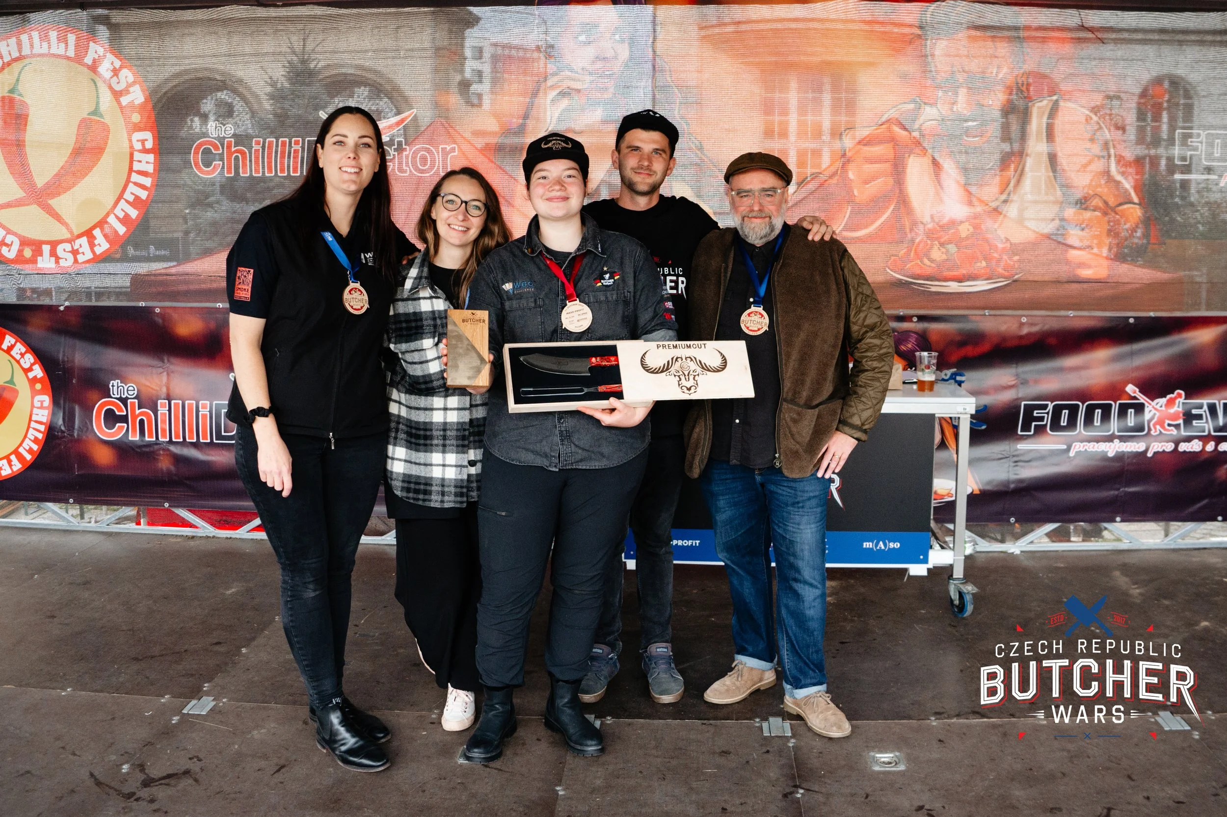 Group of five people standing together holding awards and a box, at a Czech Republic butchery competition event, with banners and logos in the background.
