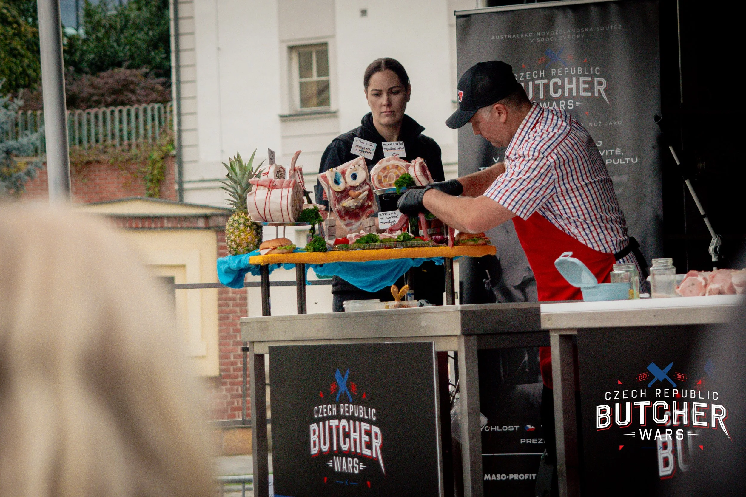 A man in a checkered shirt and red apron prepares meat at a butcher stand during an outdoor event, with a woman observing. The stand has a sign for 'Czech Republic Butcher Wars' and decorative meat sculptures, including a pineapple, on the table.