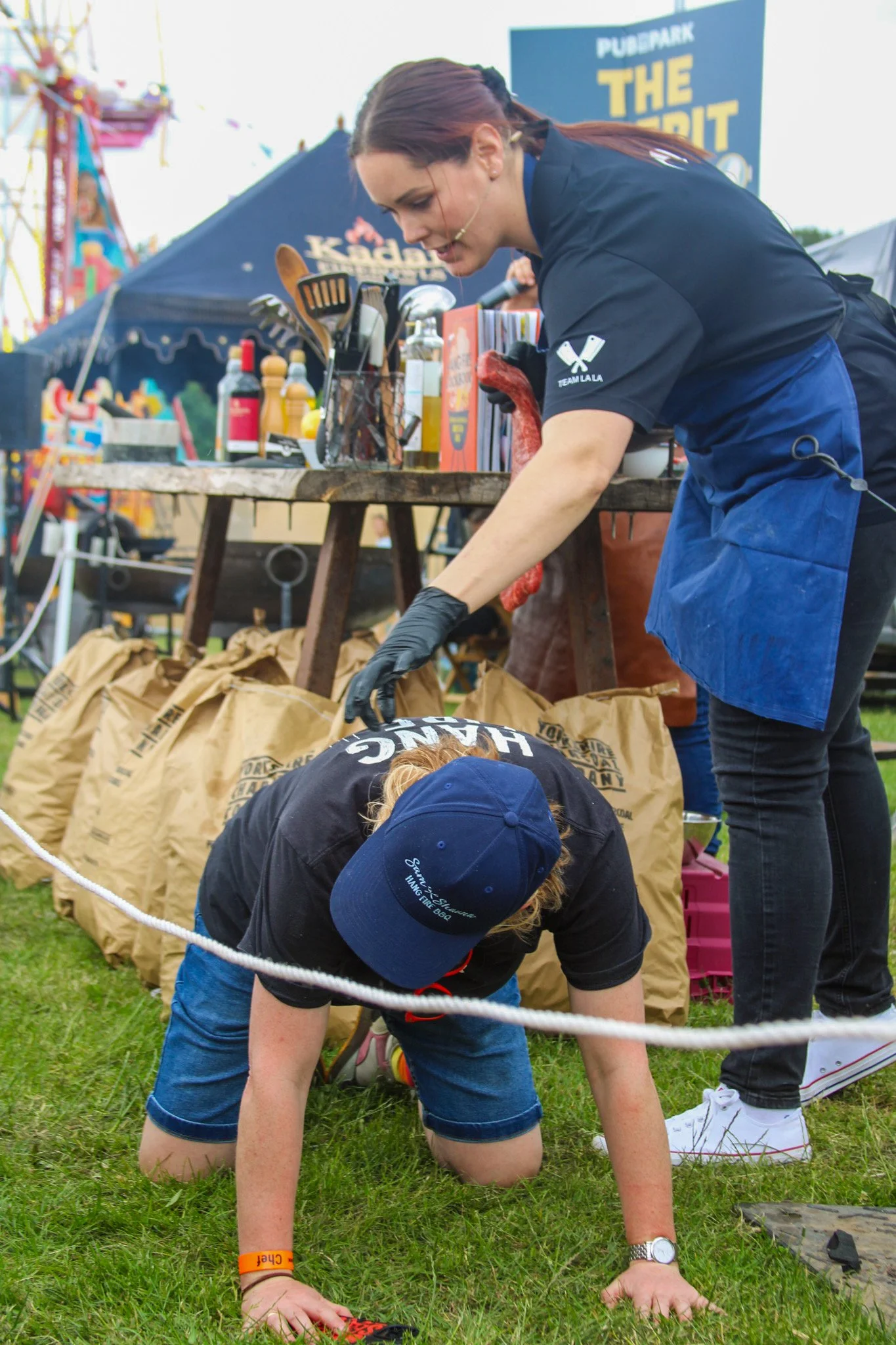 A woman in black gloves bending over a person on all fours on a grassy field at an outdoor event. The person on all fours is wearing a blue cap, black t-shirt, and denim shorts.