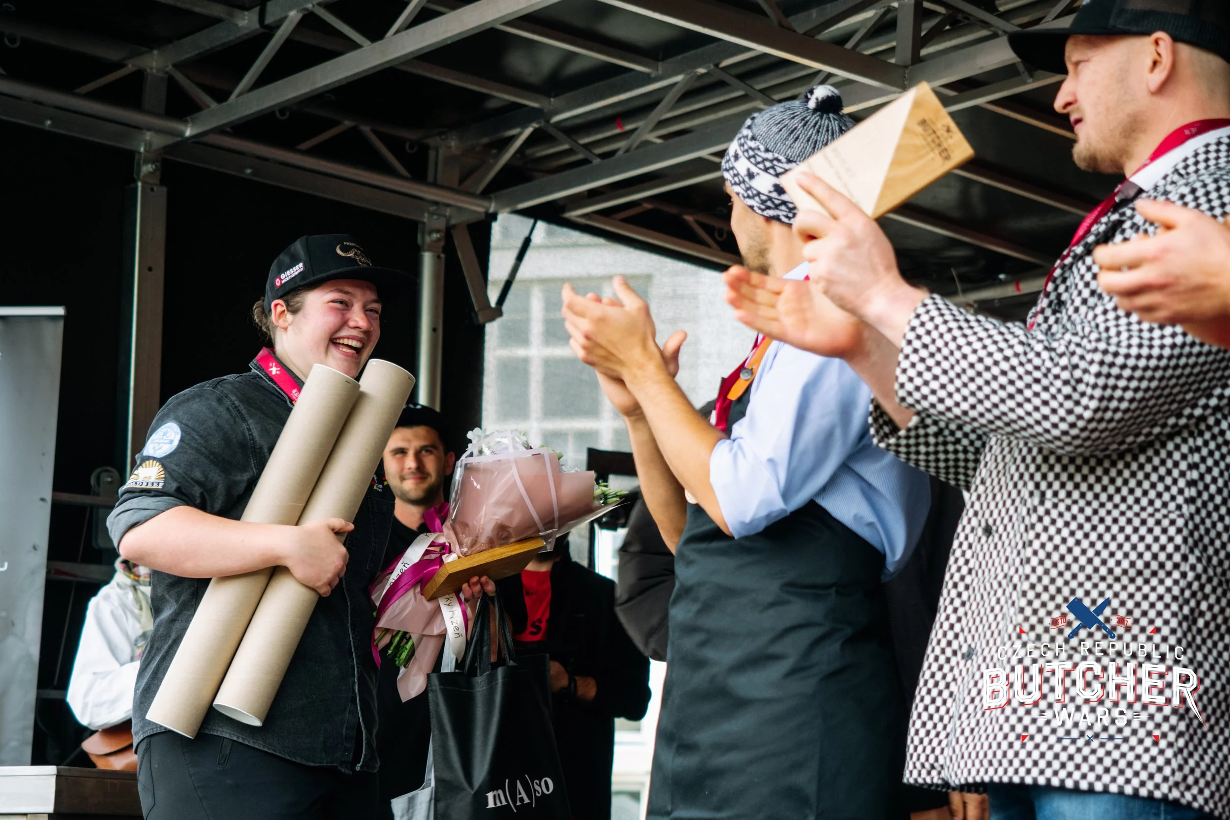 A young woman receives awards and a bouquet of flowers at a celebration, with people clapping and smiling, at an event called 'Oech Reppublic Butcher Wars'.
