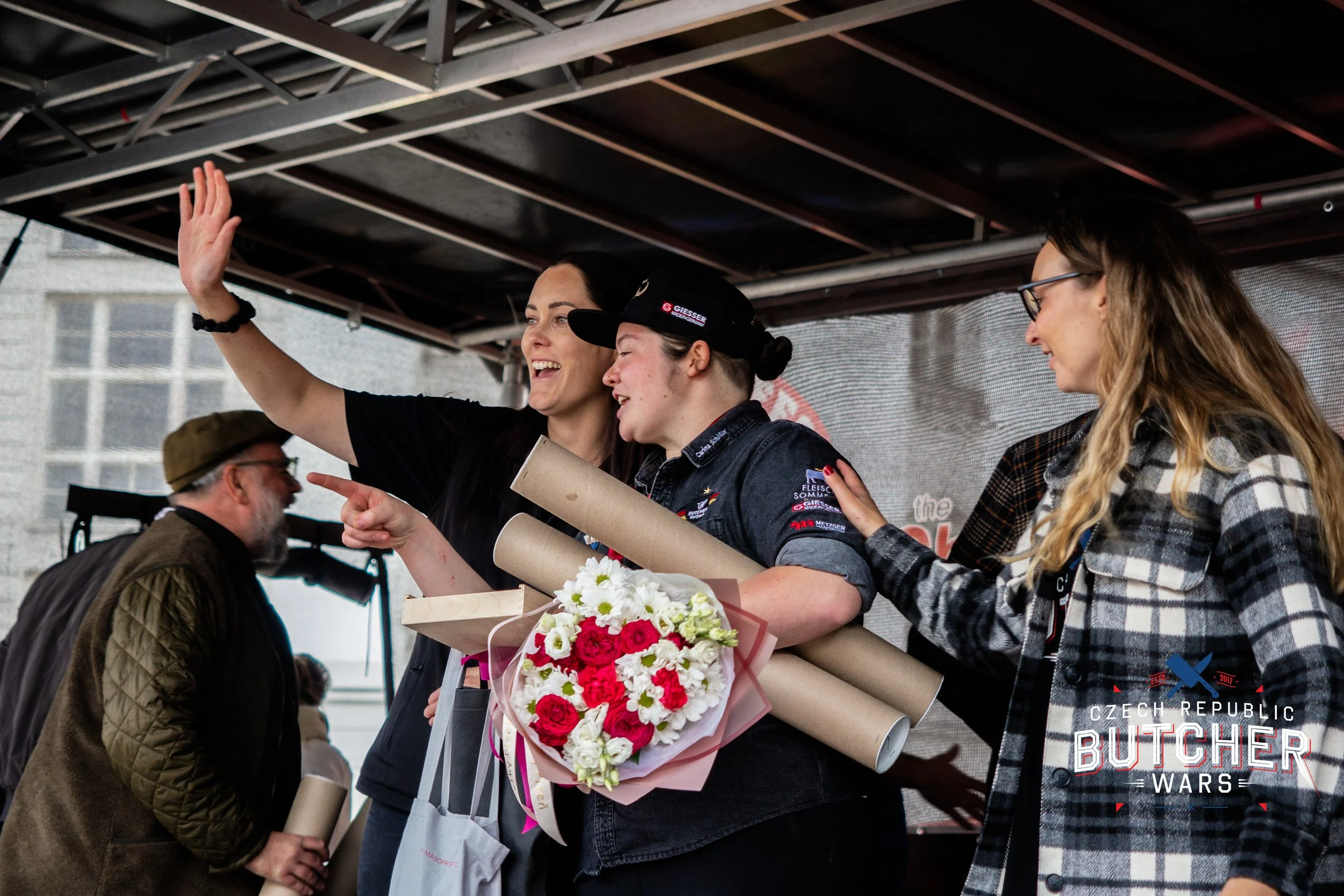 Women celebrating on stage, with one holding a bouquet of flowers and rolled-up certificates, another smiling and gesturing, and others in the background, during an awards event at Czech Republic Butcher Wars.