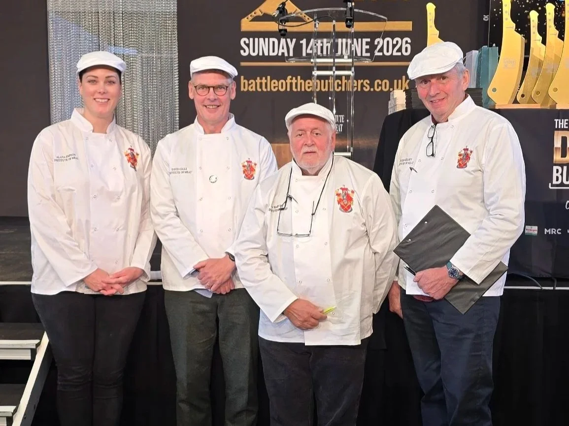 Group of four competition butchery judges in white uniforms and hats standing in front of a black background with event details and trophies on display.