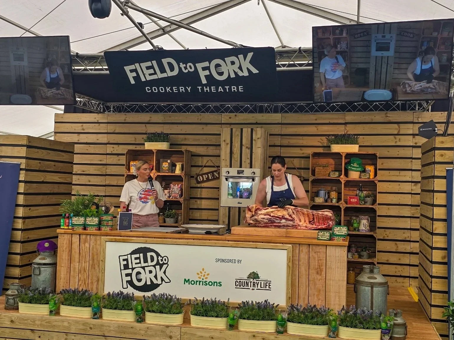 A butchery demonstration at Field to Fork Cookery Theatre with two women preparing meat on a wooden stage, decorated with wood paneling, shelves with products and plants, and large display screens showing the scene.