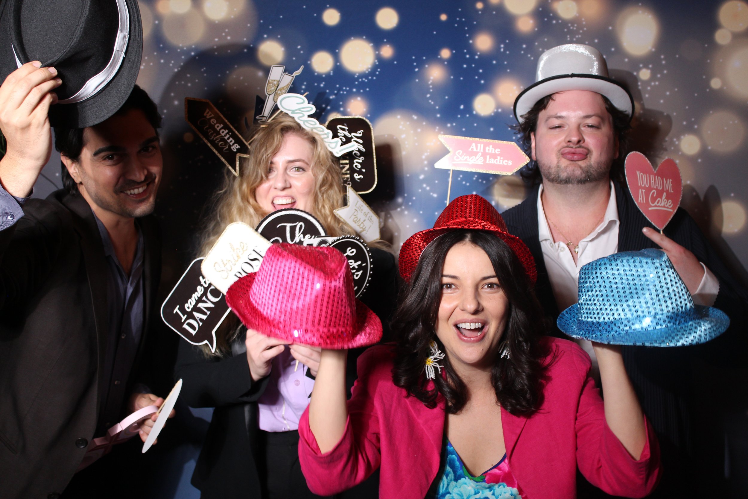 A woman DJ with black hair and wearing a black dress, using DJ equipment at a party. In the background, through a photo booth cutout, three women are holding drinks, wearing festive accessories, and smiling with a "PARTY" sign, surrounded by warm, festive lighting.