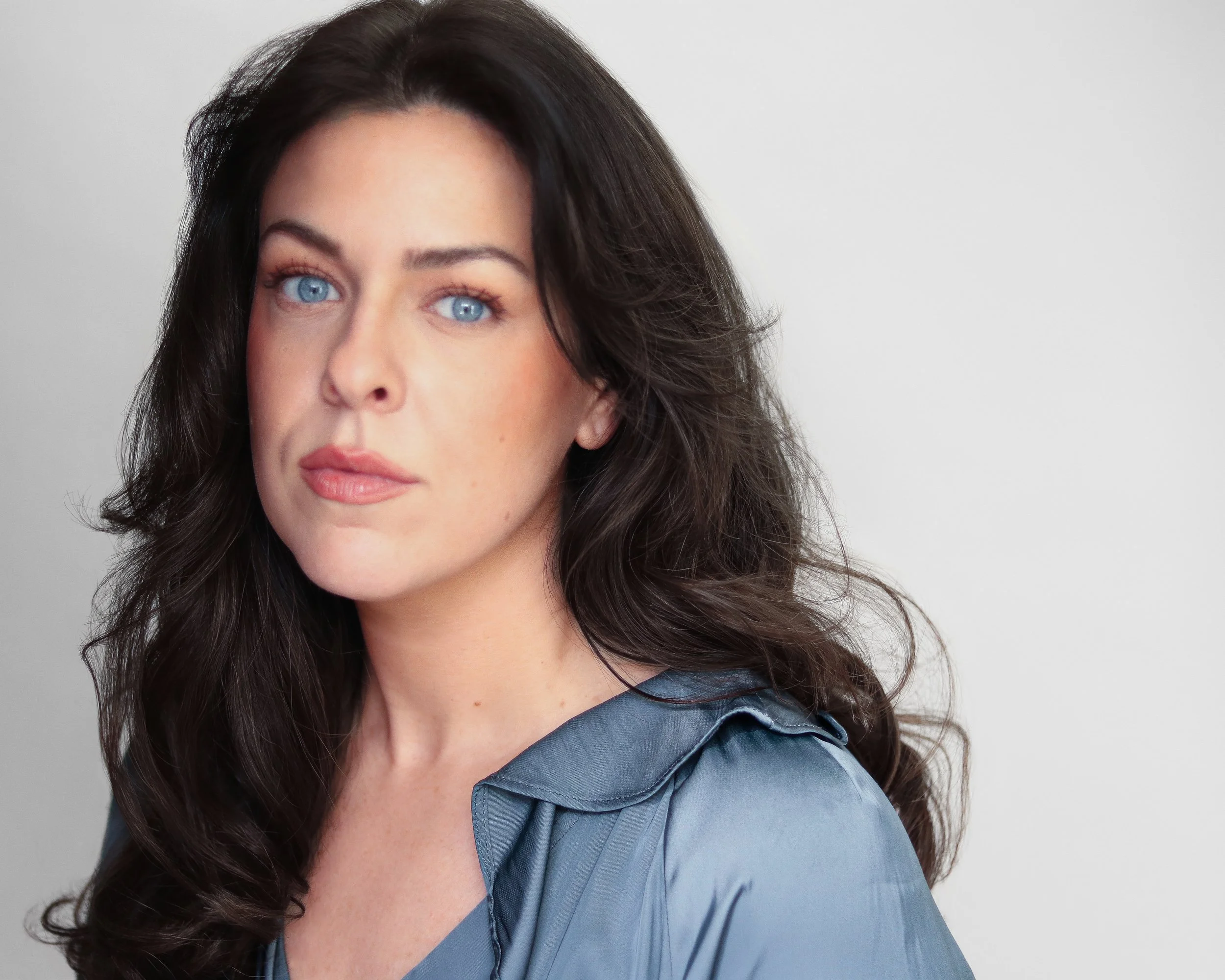 A woman with long dark hair, blue eyes, and light makeup, wearing a silky blue-gray top, posing against a plain white background.