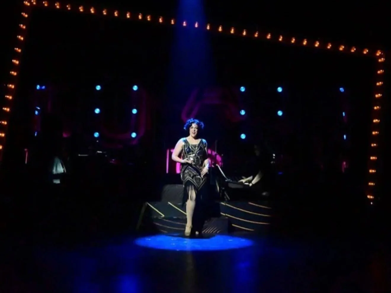 A woman in a vintage dress standing on a stage in a theater, with a microphone in hand, illuminated by a spotlight surrounded by stage lights and decorative borders, with a pianist playing behind her.