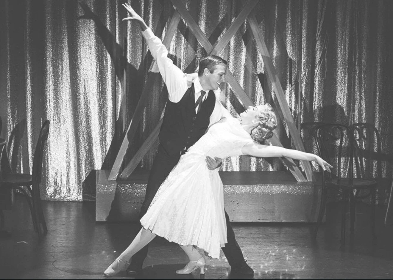 A vintage black-and-white photograph of a man and a woman dancing together on a stage with a decorated backdrop and chairs. The woman wears a light-colored dress and the man wears a dark vest and trousers. They appear to be performing a dance routine.