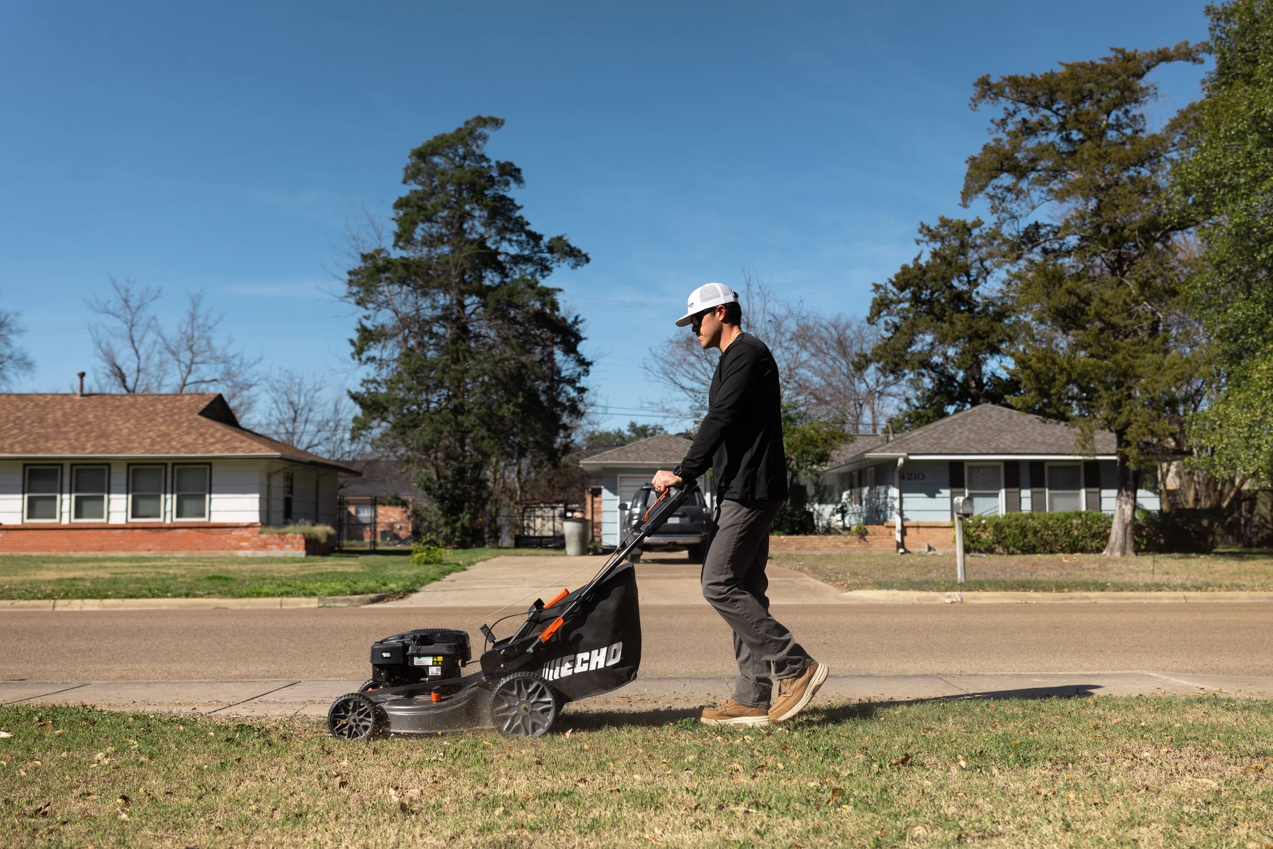 A man mowing the lawn with a lawnmower on a suburban street during daytime.