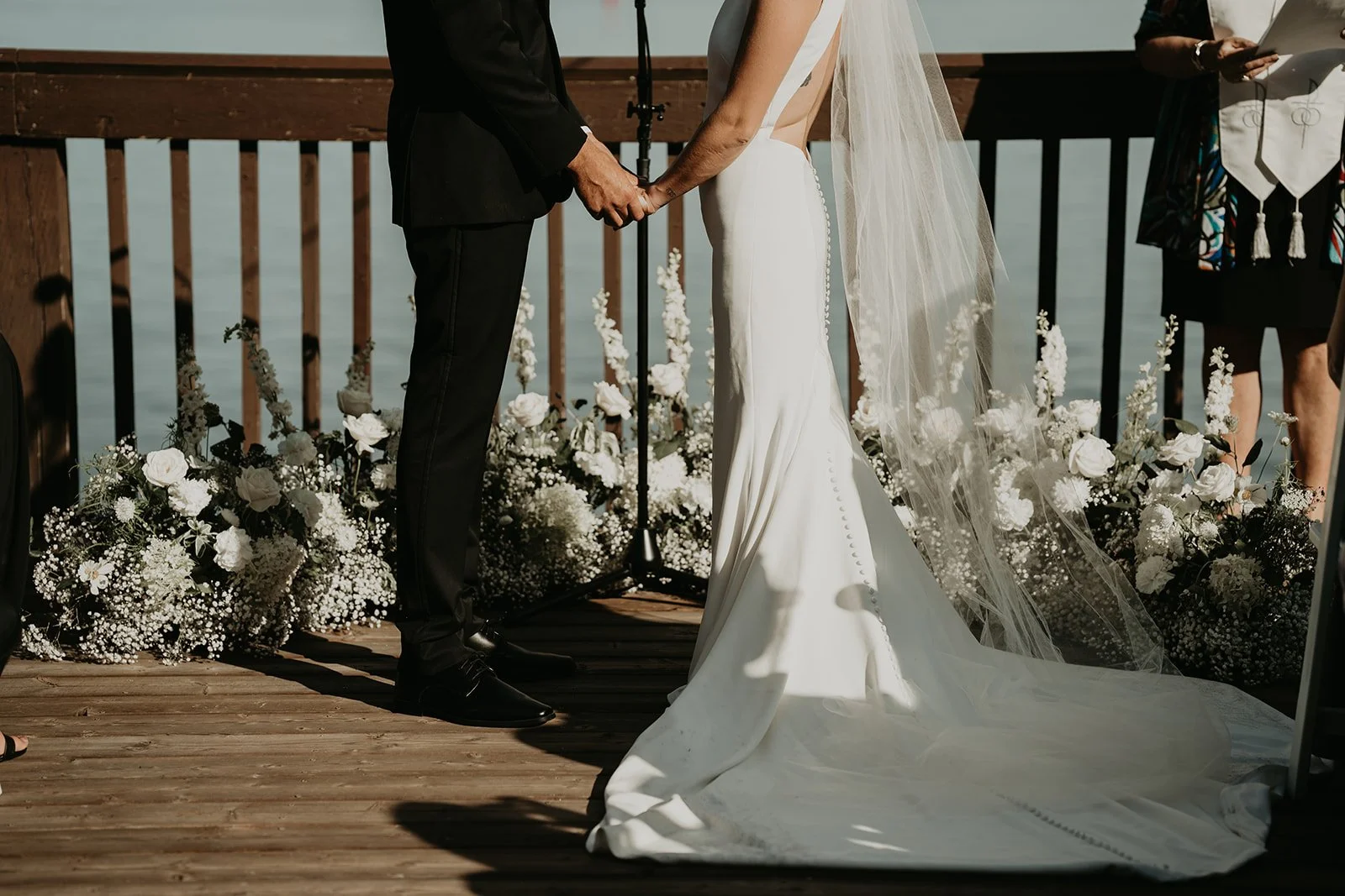 A bride and groom holding hands during their wedding ceremony on a wooden dock, with a background of water and floral arrangements.