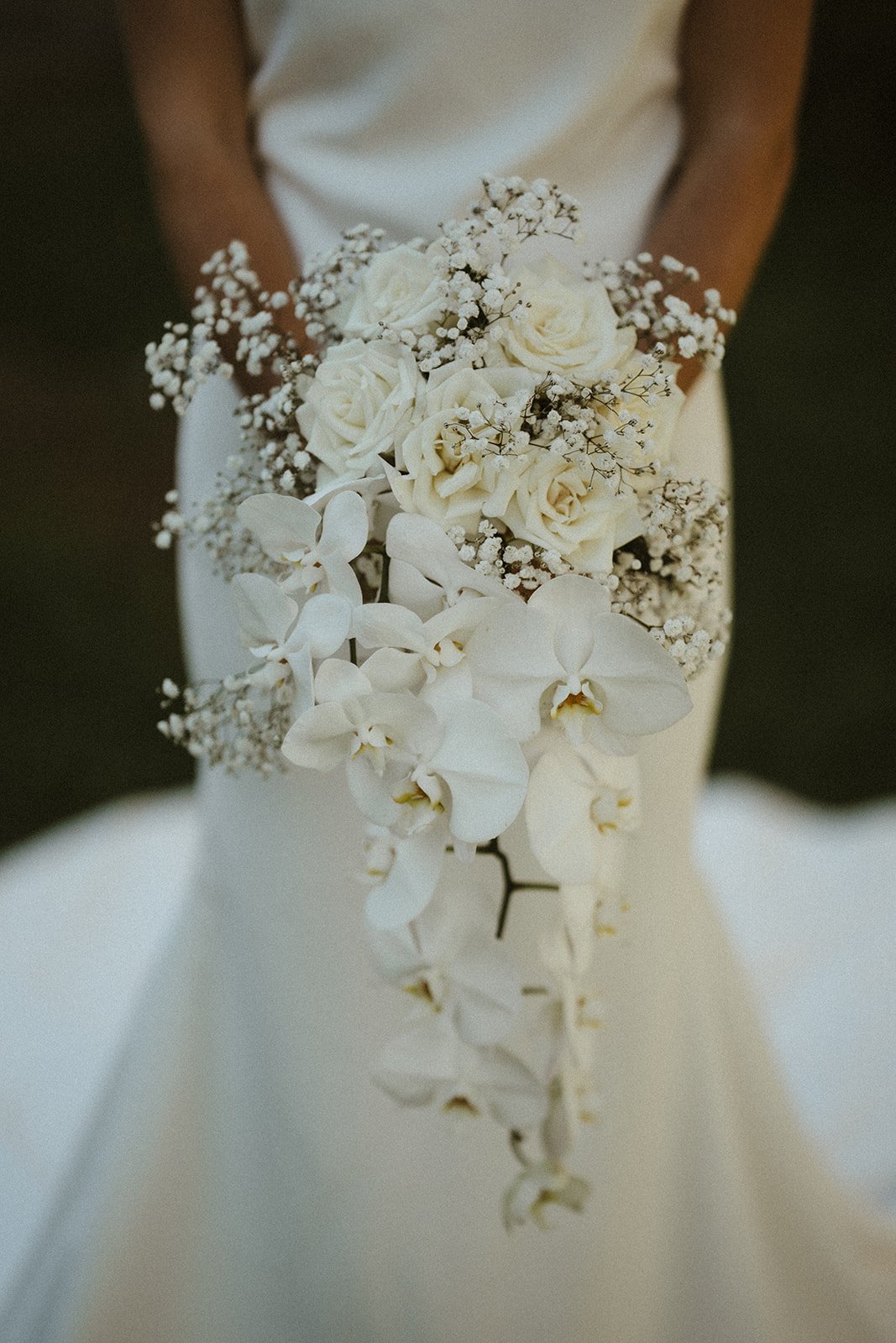 Close-up of a bride holding a cascading bouquet of white roses, orchids, and baby's breath, with a white wedding dress in the background.