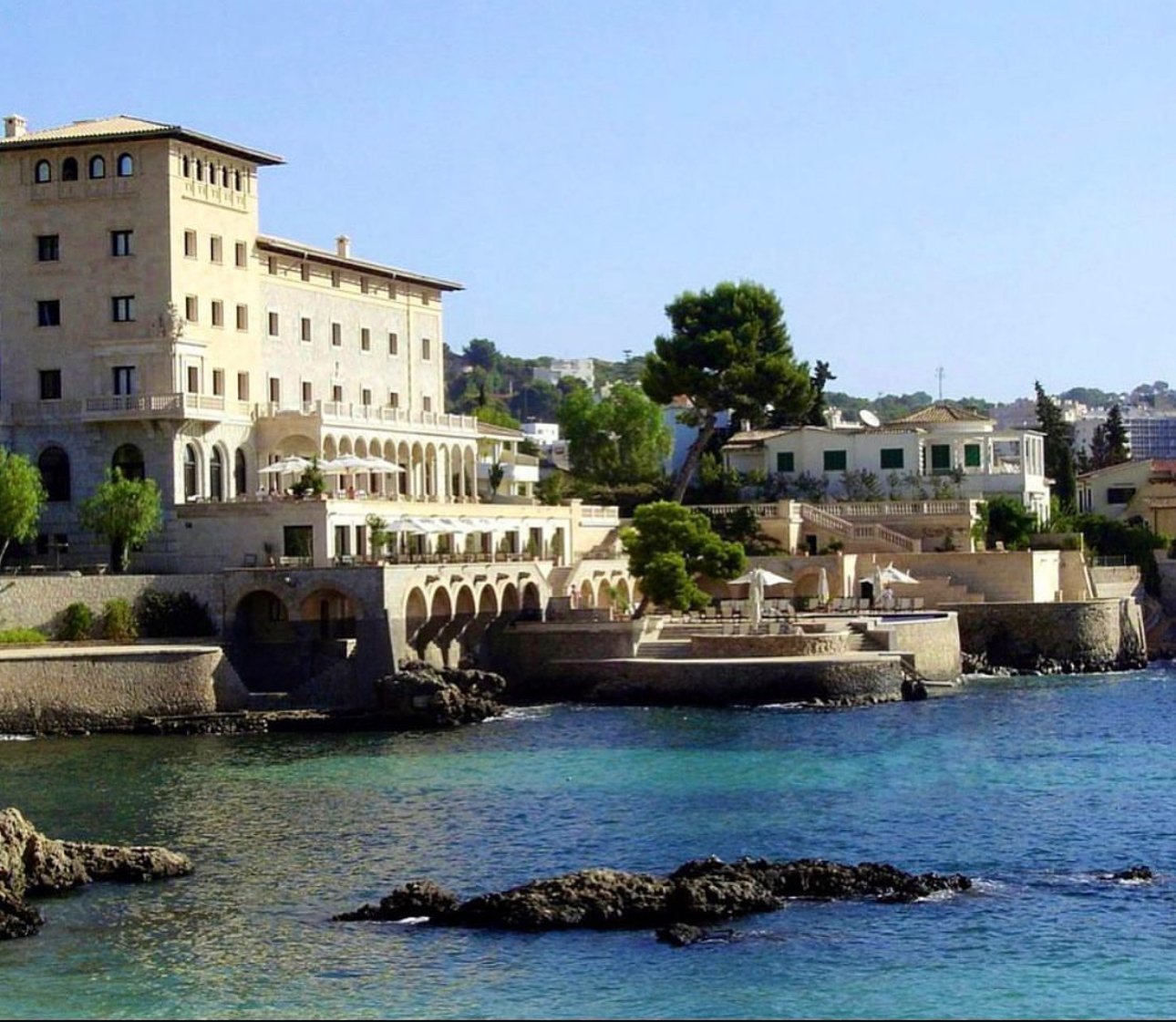 Luxurious waterfront villa with multiple terraces and arches, surrounded by lush trees and overlooking calm blue water.