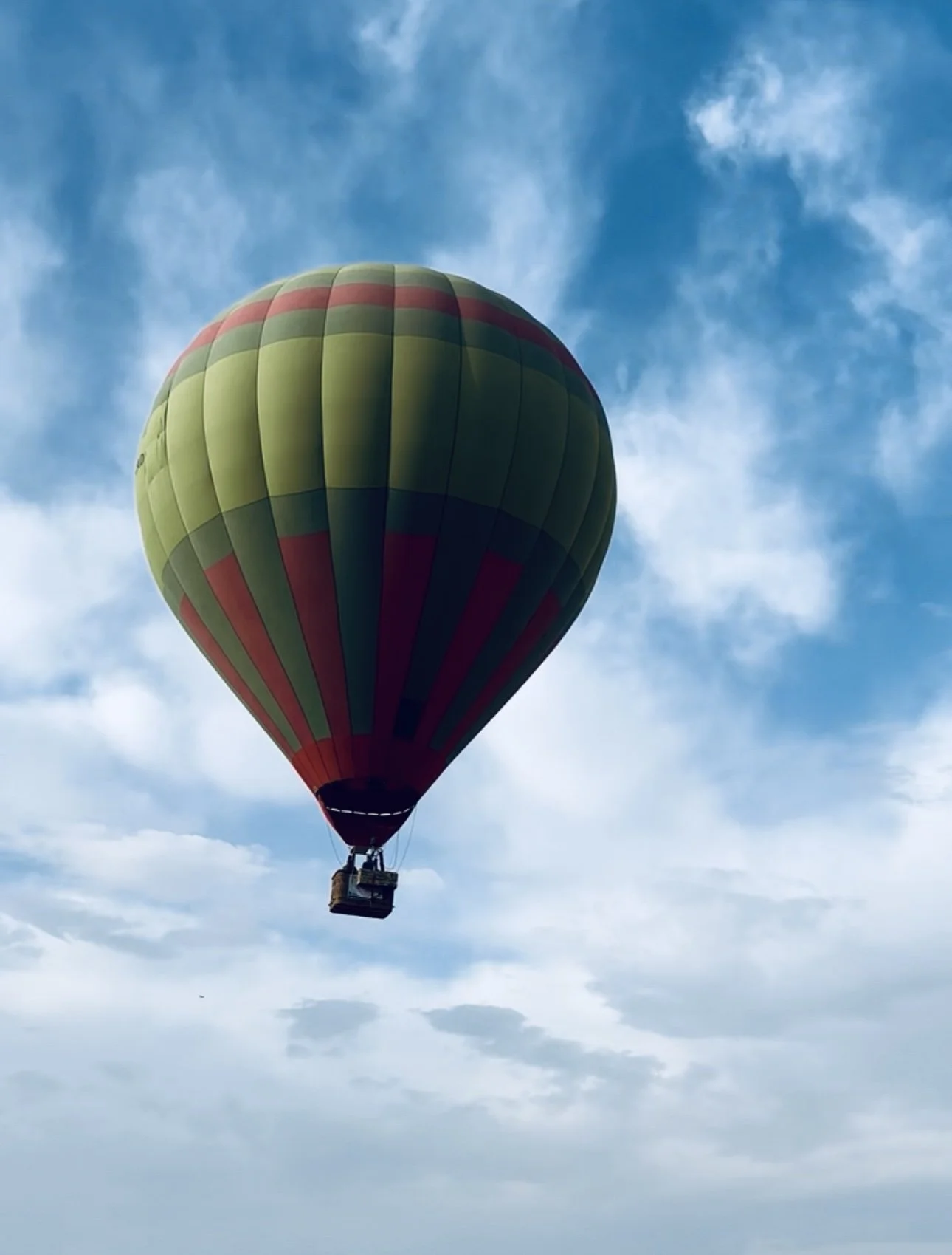 A hot air balloon with green, red, and dark panels floating in a blue sky with scattered clouds.
