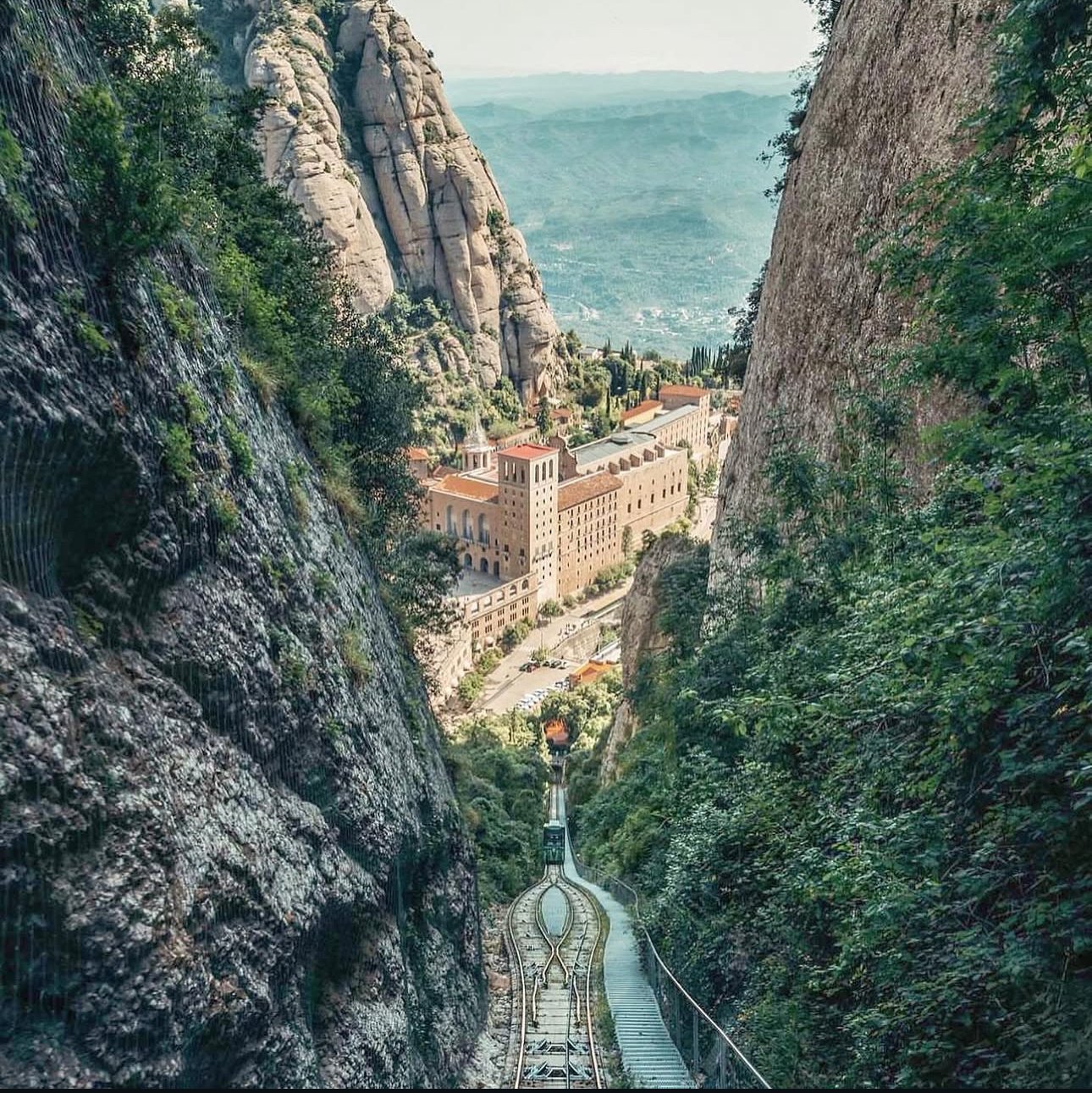 A funicular railway descending between steep rocky cliffs surrounded by greenery, leading to a large building with towers and red roofs in a valley with a cityscape in the background.