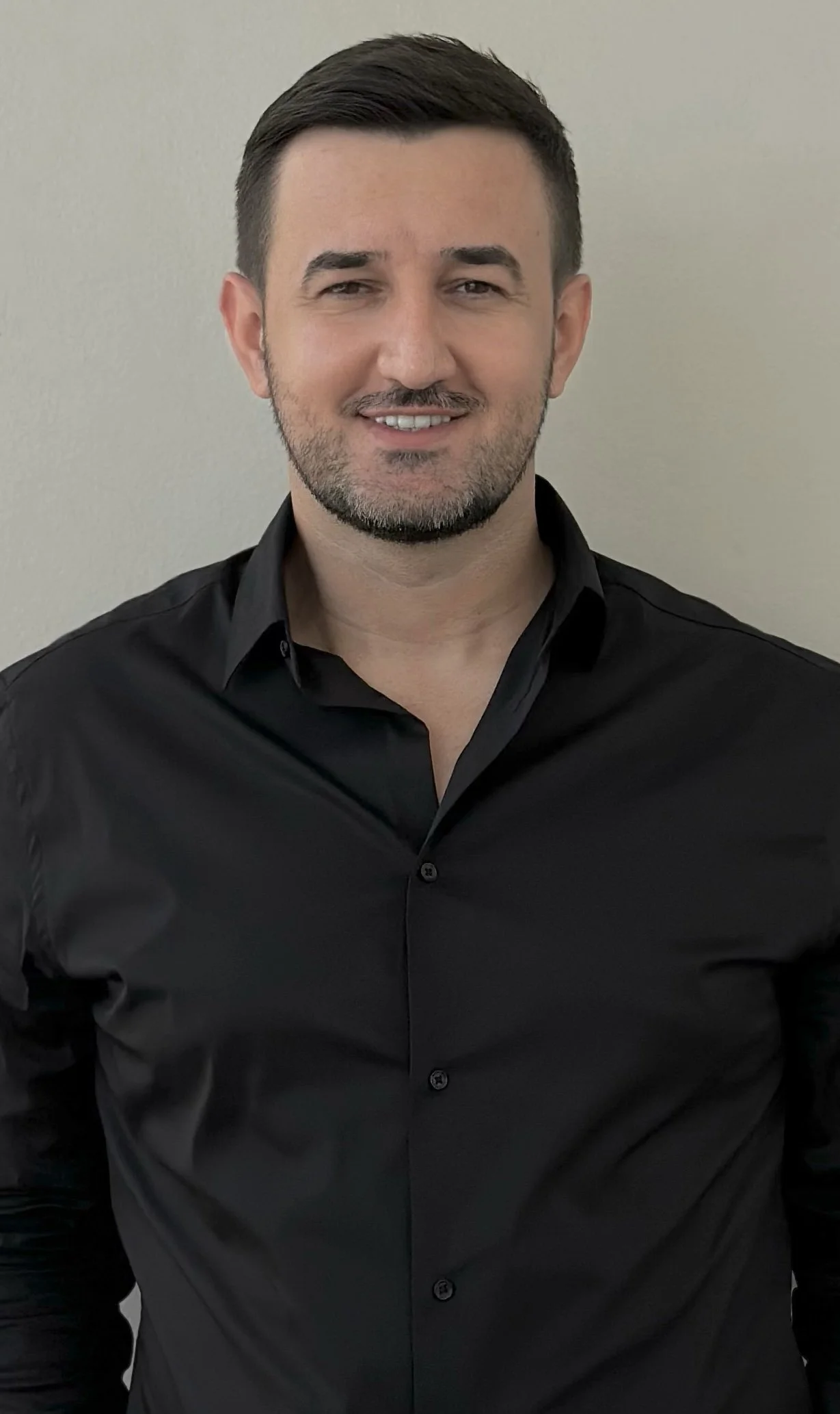 A man with short dark hair and a trimmed beard, dressed in a black button-up shirt, standing against a light-colored plain wall, smiling and looking at the camera.