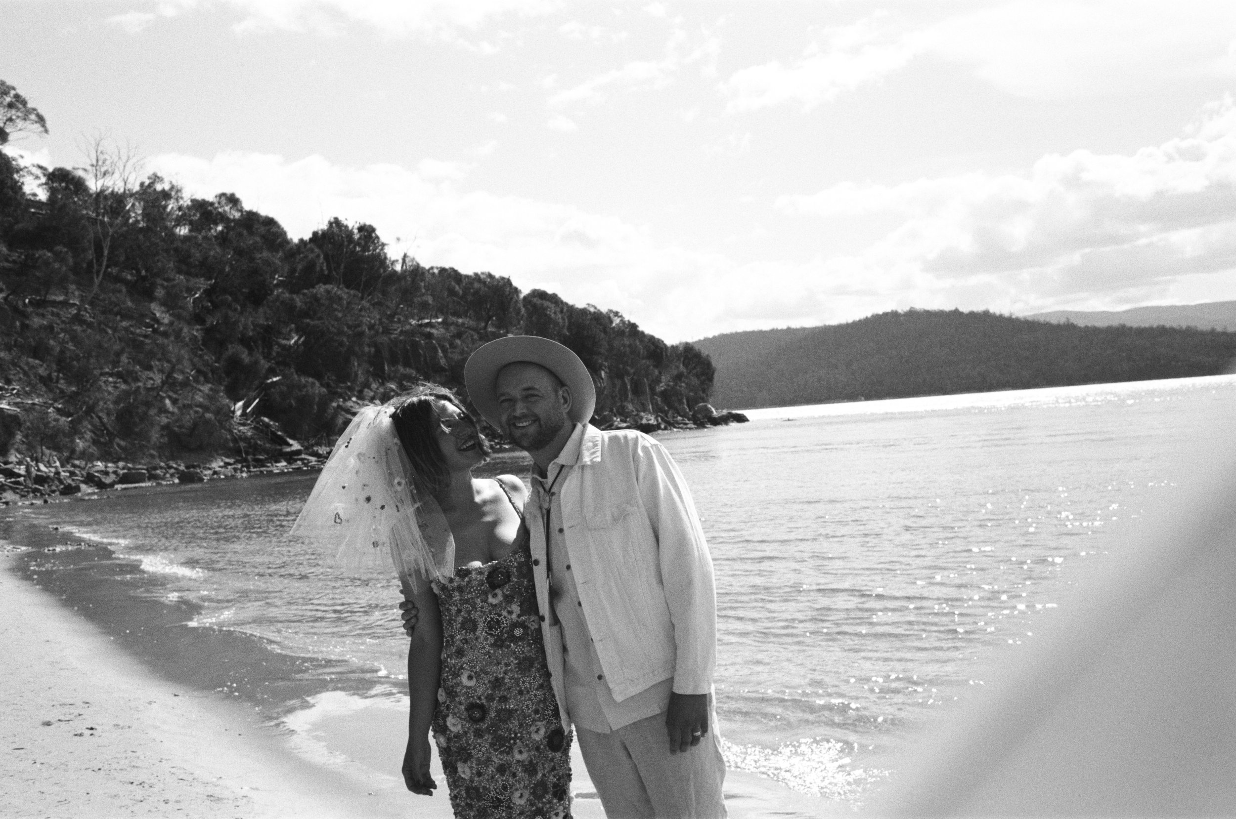 A couple in wedding attire standing on a beach by the water, smiling and embracing, with trees and distant hills in the background.