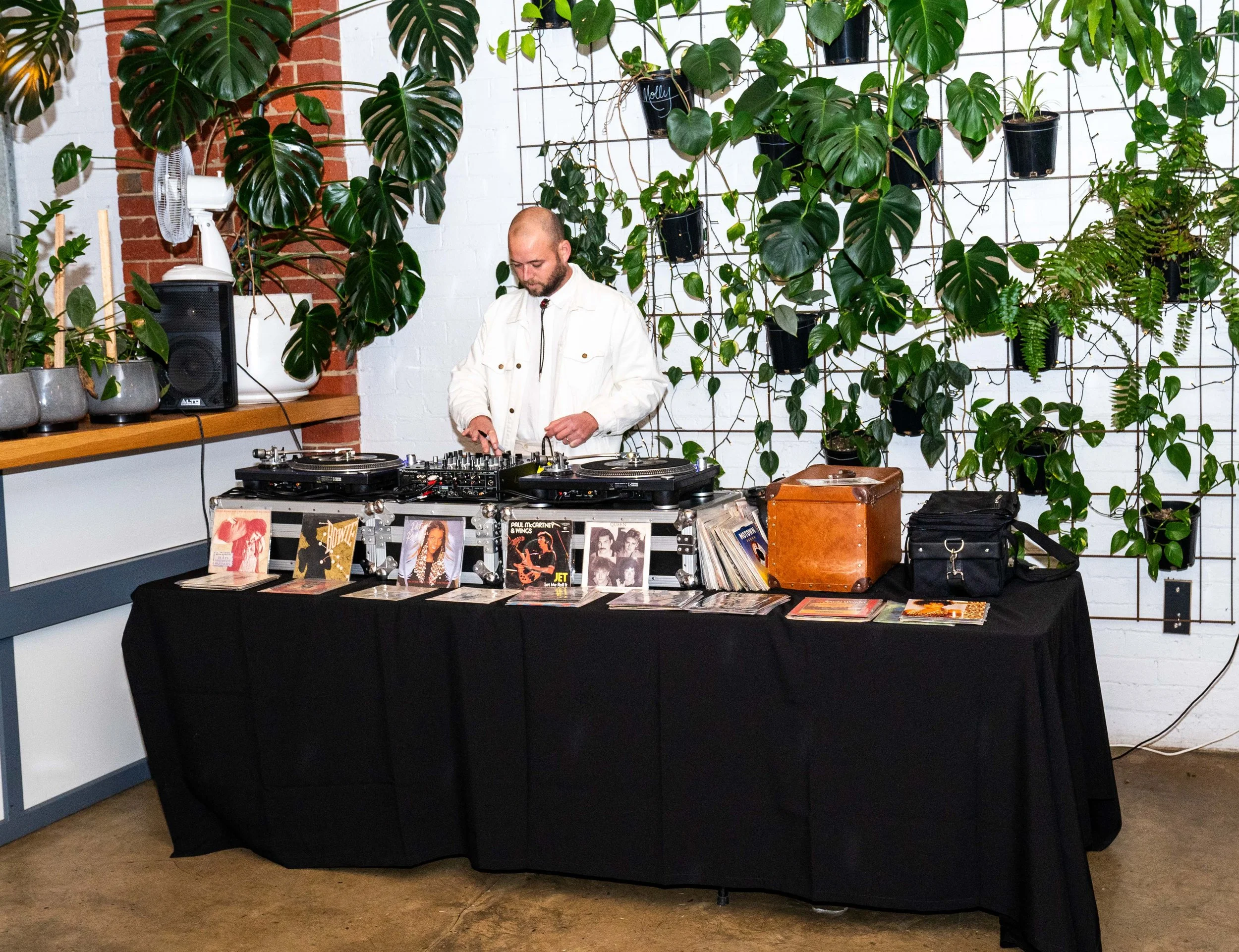 A DJ stands behind a table with vinyl records and turntables, in a room decorated with various green plants on a wire grid wall.