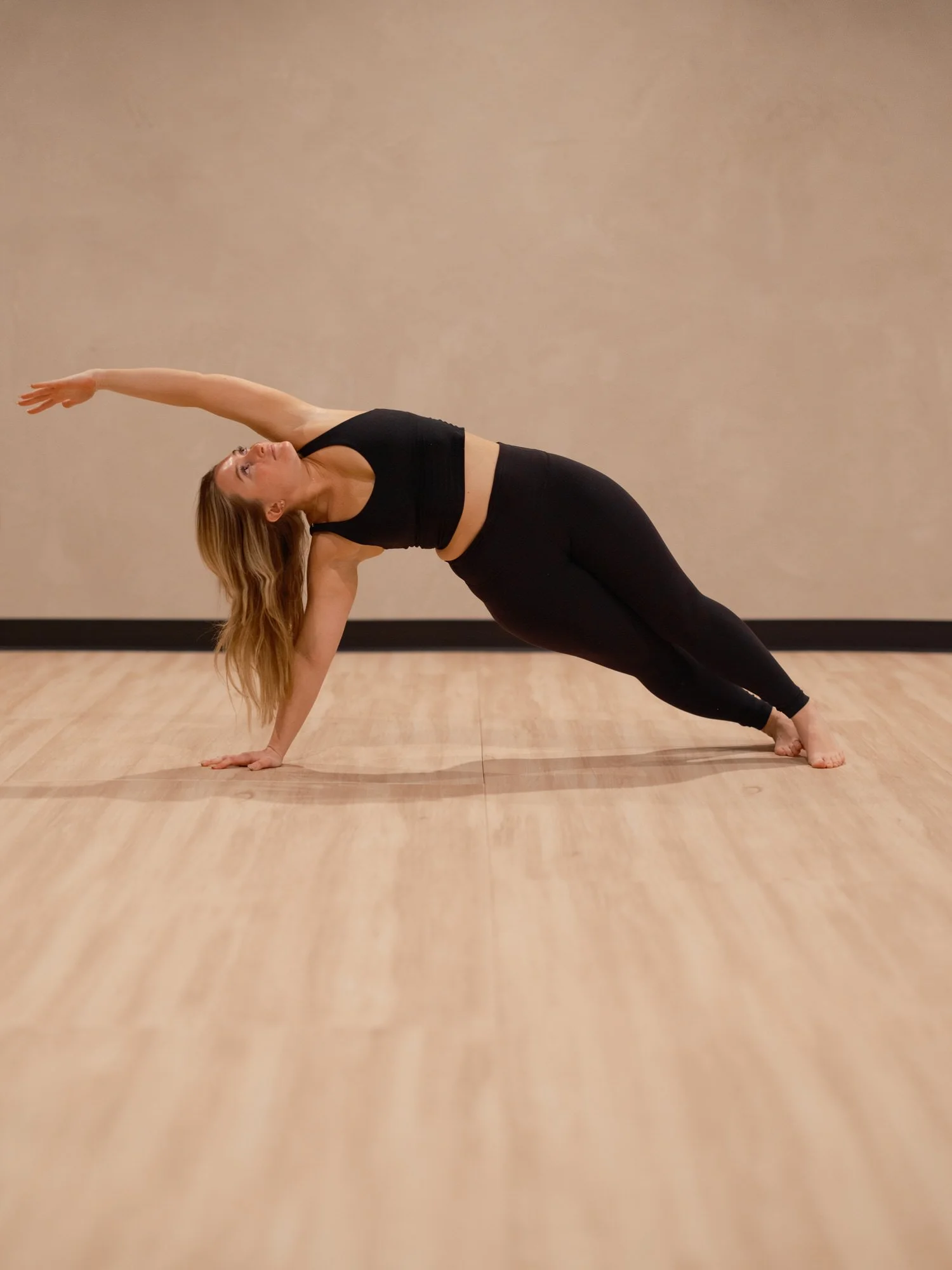 A woman in black workout clothes doing a yoga side plank pose on a wooden floor against a plain wall in 8th State Hot Yoga studio.