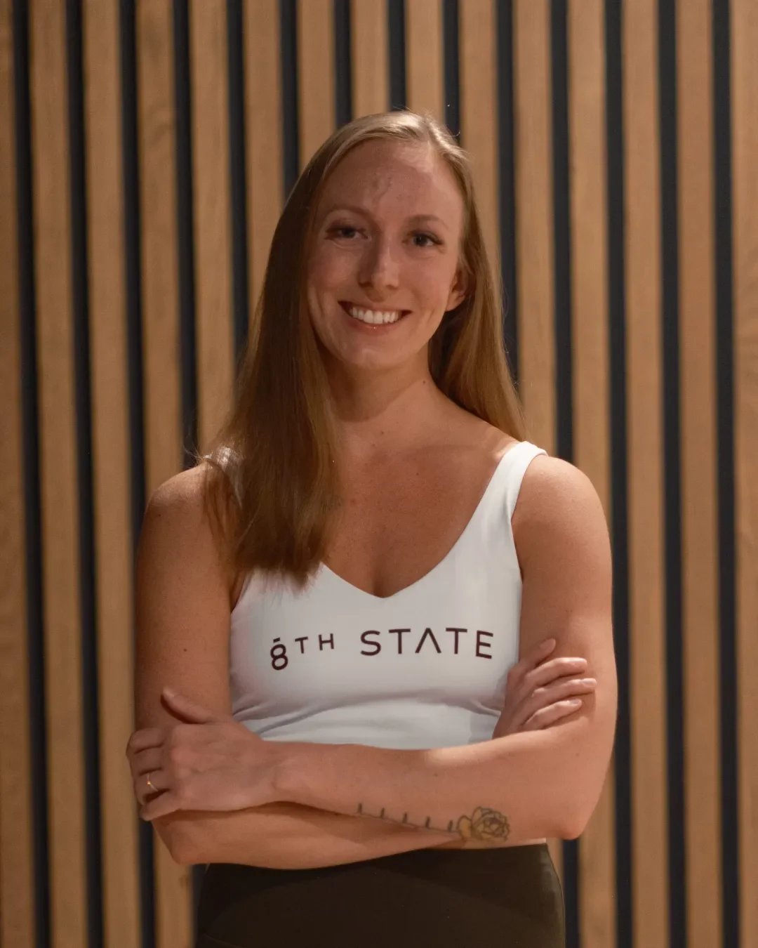 A young woman with long red hair, smiling with arms crossed, wearing a white tank top that says '8TH STATE' in front of a wooden slat background.