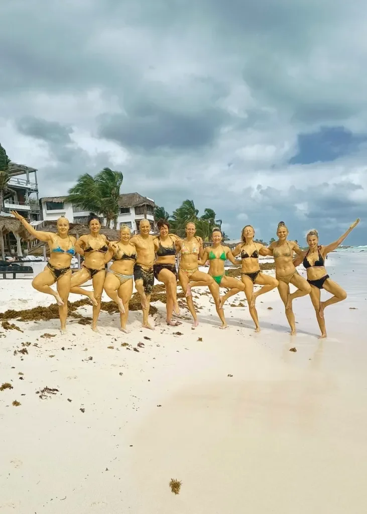 A group of women in swimsuits covered in sand, standing on a beach with their arms around each other, under a cloudy sky, with palm trees and beach houses in the background.