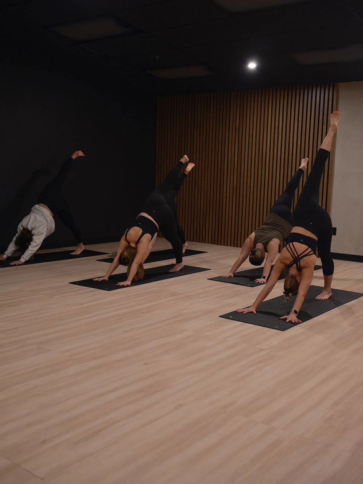 Four people practicing yoga at 8th State Hot Yoga studio, performing split downward dog poses on black yoga mats.