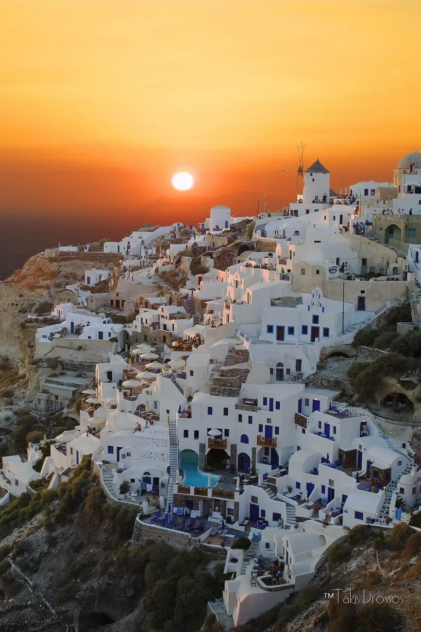 Sunset over the white buildings of Oia, Santorini, with a windmill on a hillside and the sea in the background.