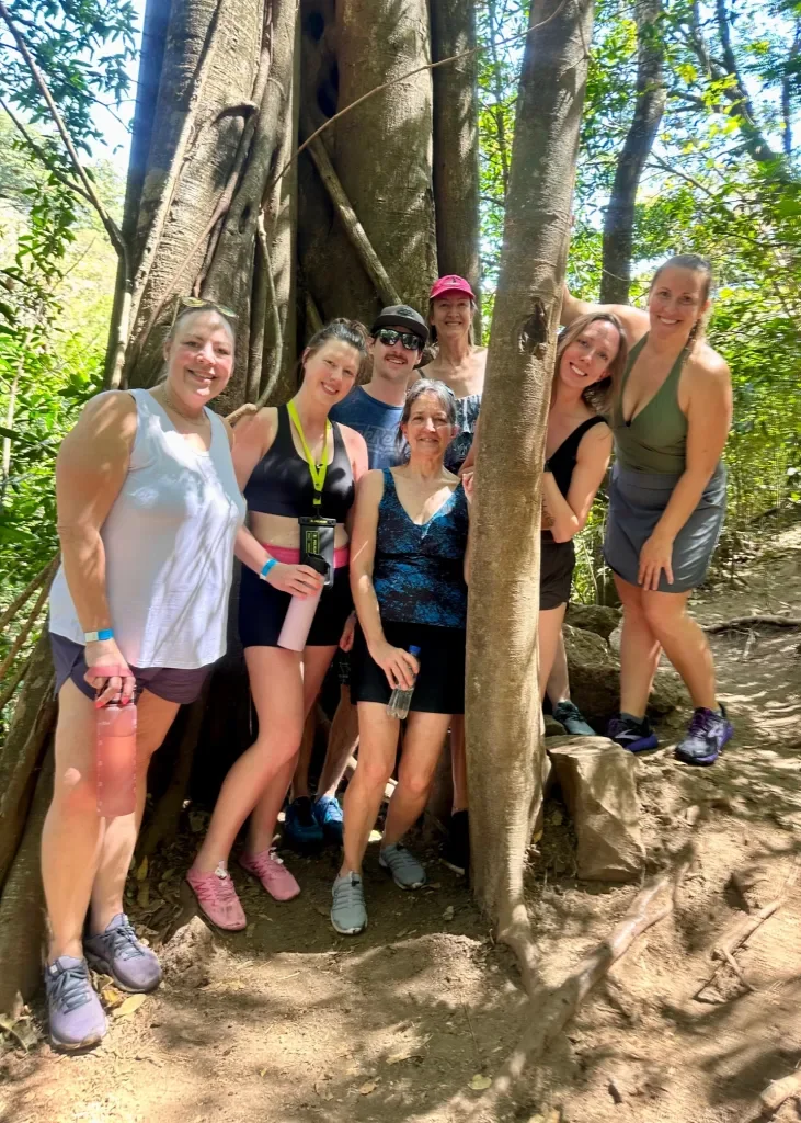 Group of seven people posing outdoors near a large tree in a forest, smiling and dressed in activewear.